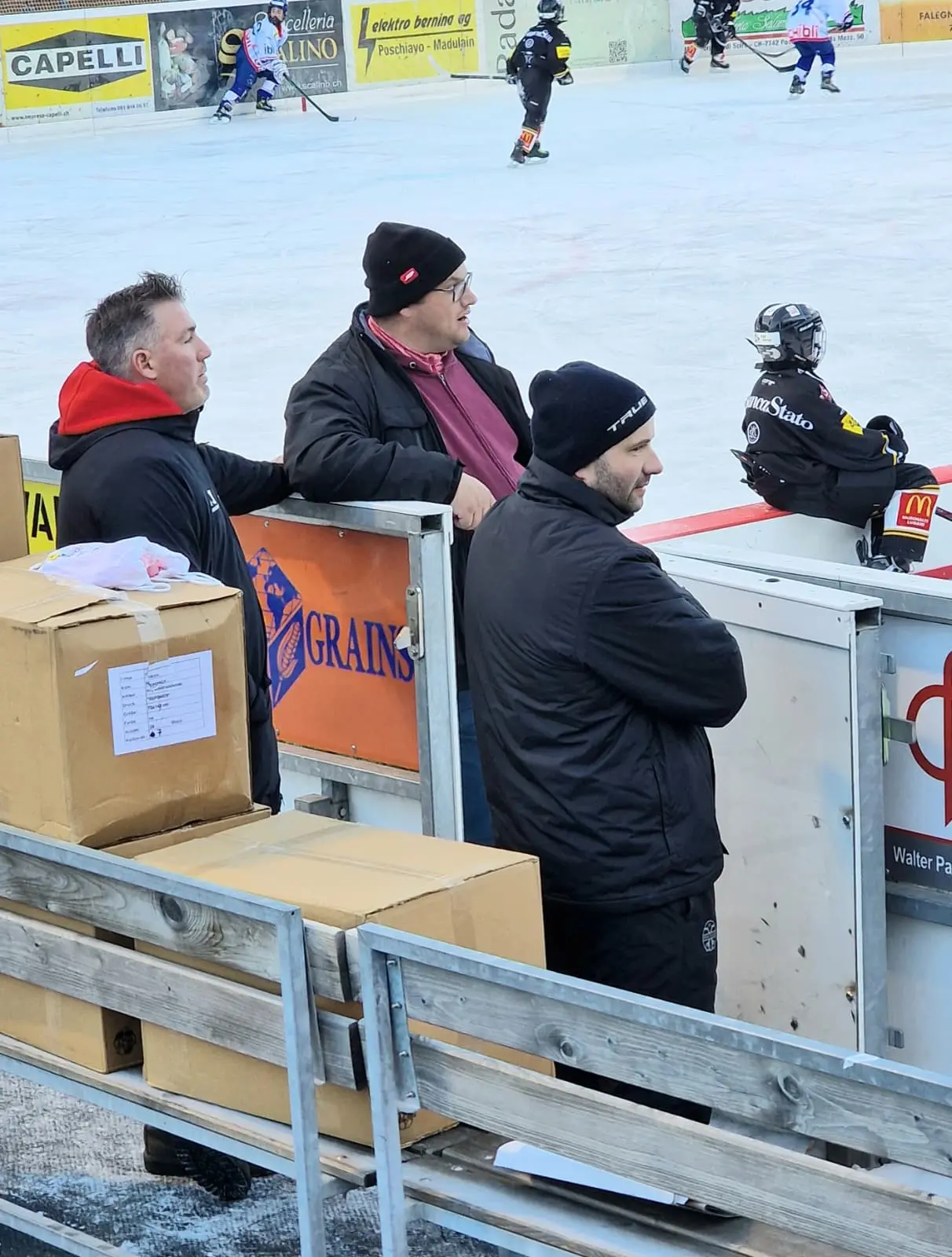 Tre uomini in abbigliamento invernale osservano una partita di hockey su ghiaccio, mentre si trovano accanto a una barriera. Sullo sfondo, si vedono gioc