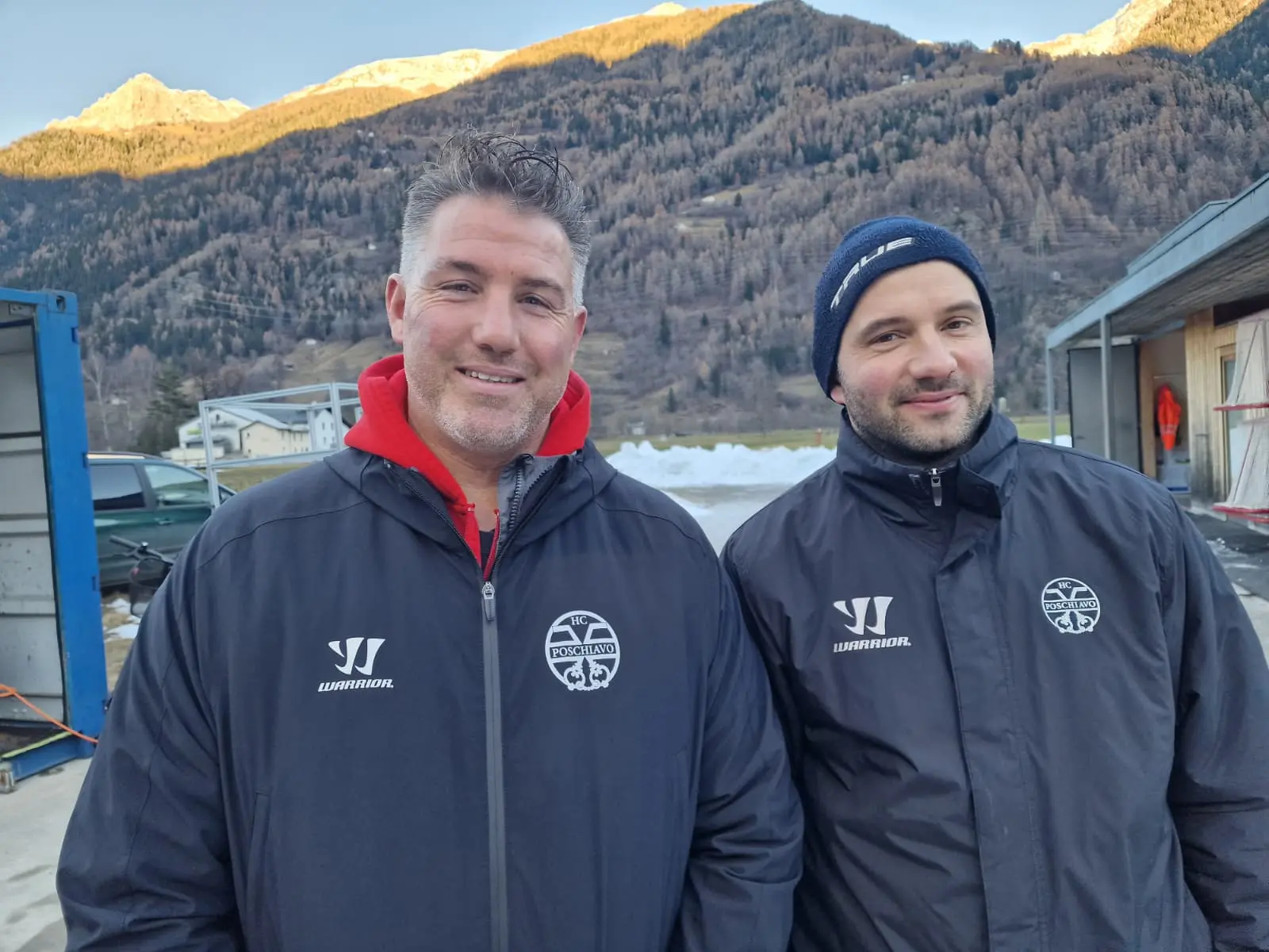 Due uomini sorridenti indossano giacche nere con loghi sportivi, mentre si trovano all'aperto con montagne sullo sfondo. La luce del sole illumina la scena,