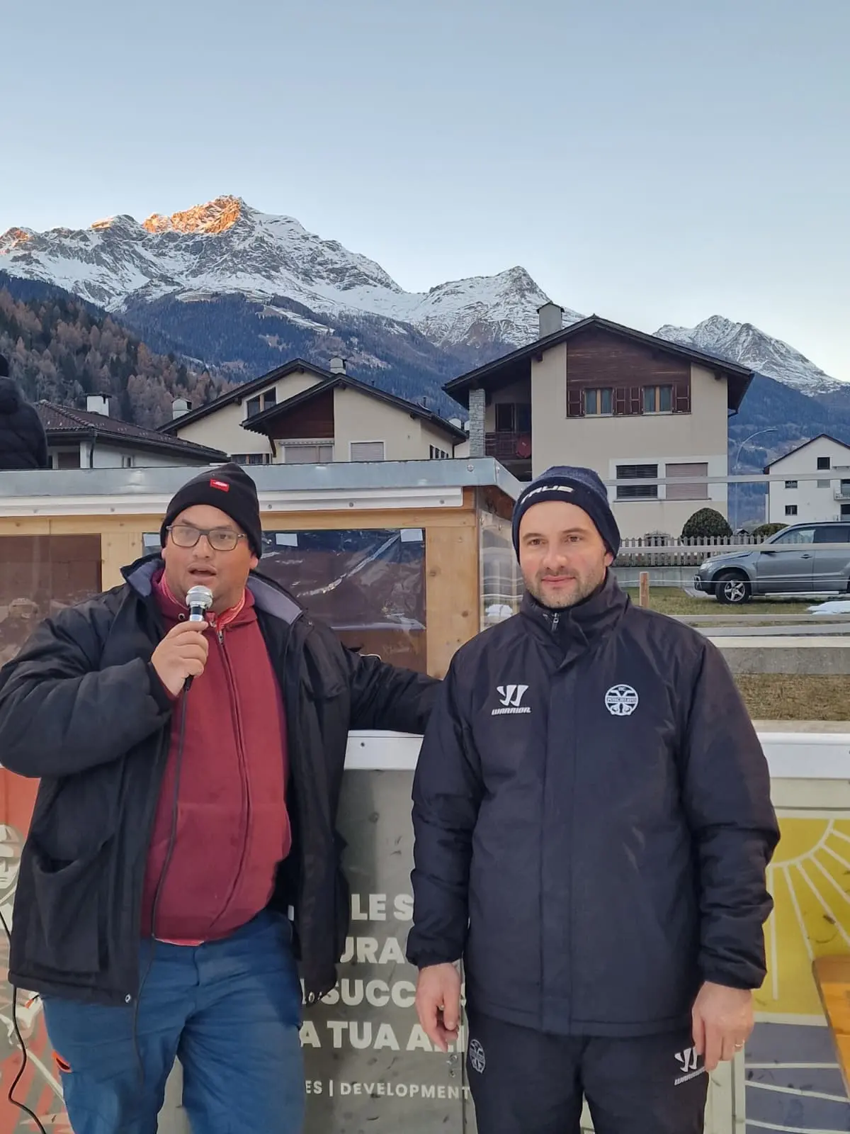 Due uomini stanno parlando in un ambiente all'aperto. Uno tiene un microfono e indossa un giubbotto nero con il logo di una squadra. L'altro indossa un ab