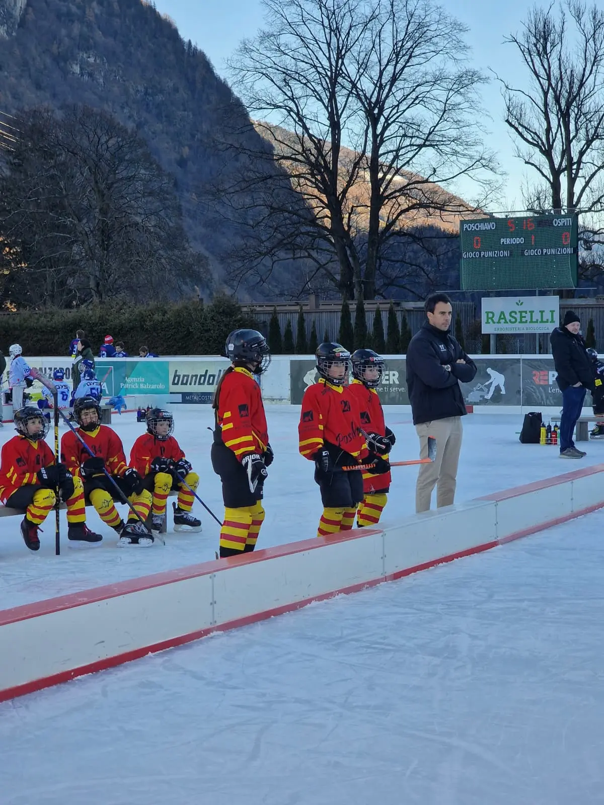 Gruppo di giovani giocatori di hockey su ghiaccio, vestiti con maglie rosse e gialle, in attesa sulla panchina di un campo ghiacciato. Alle sp