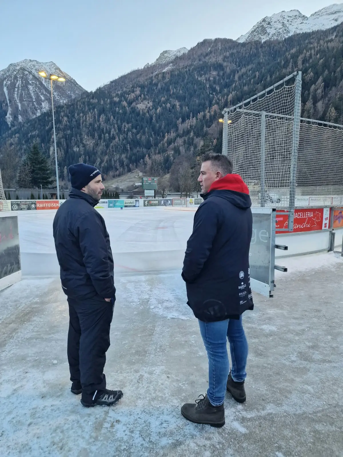 Due uomini in abbigliamento invernale stanno conversando su un campo di ghiaccio di hockey, circondati da montagne e alberi. Il cielo è sereno e chiaro.