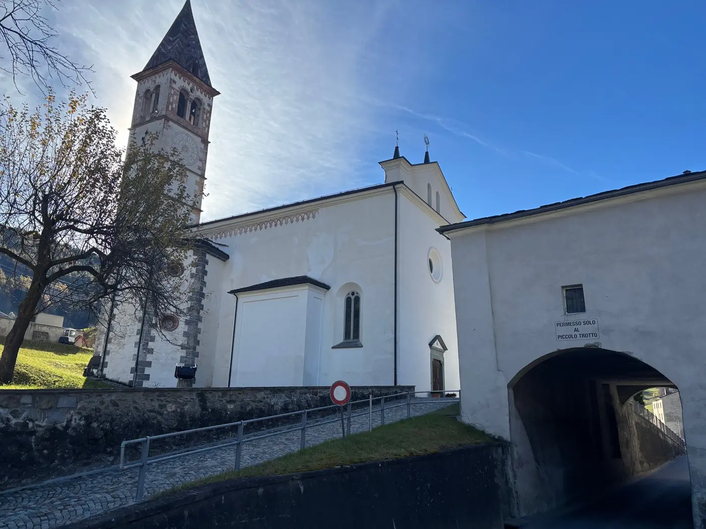 Chiesa bianca con campanile e struttura adiacente, immersa in un paesaggio sereno e soleggiato. Un sentiero di ciottoli conduce all'edificio.