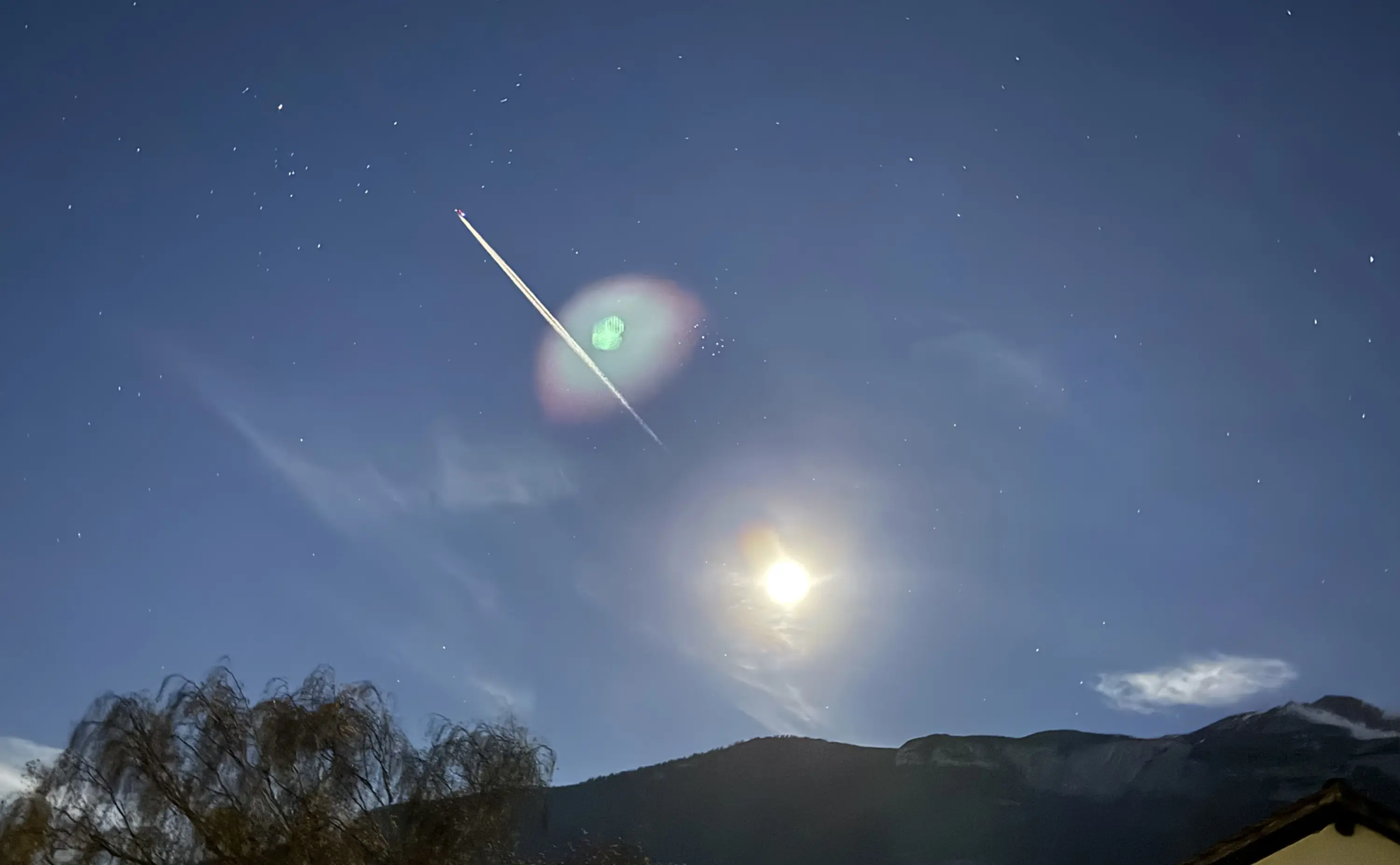 Cielo notturno stellato con una scia di un aereo e una luna luminosa. Sullo sfondo, montagne silhouette e nuvole leggere.