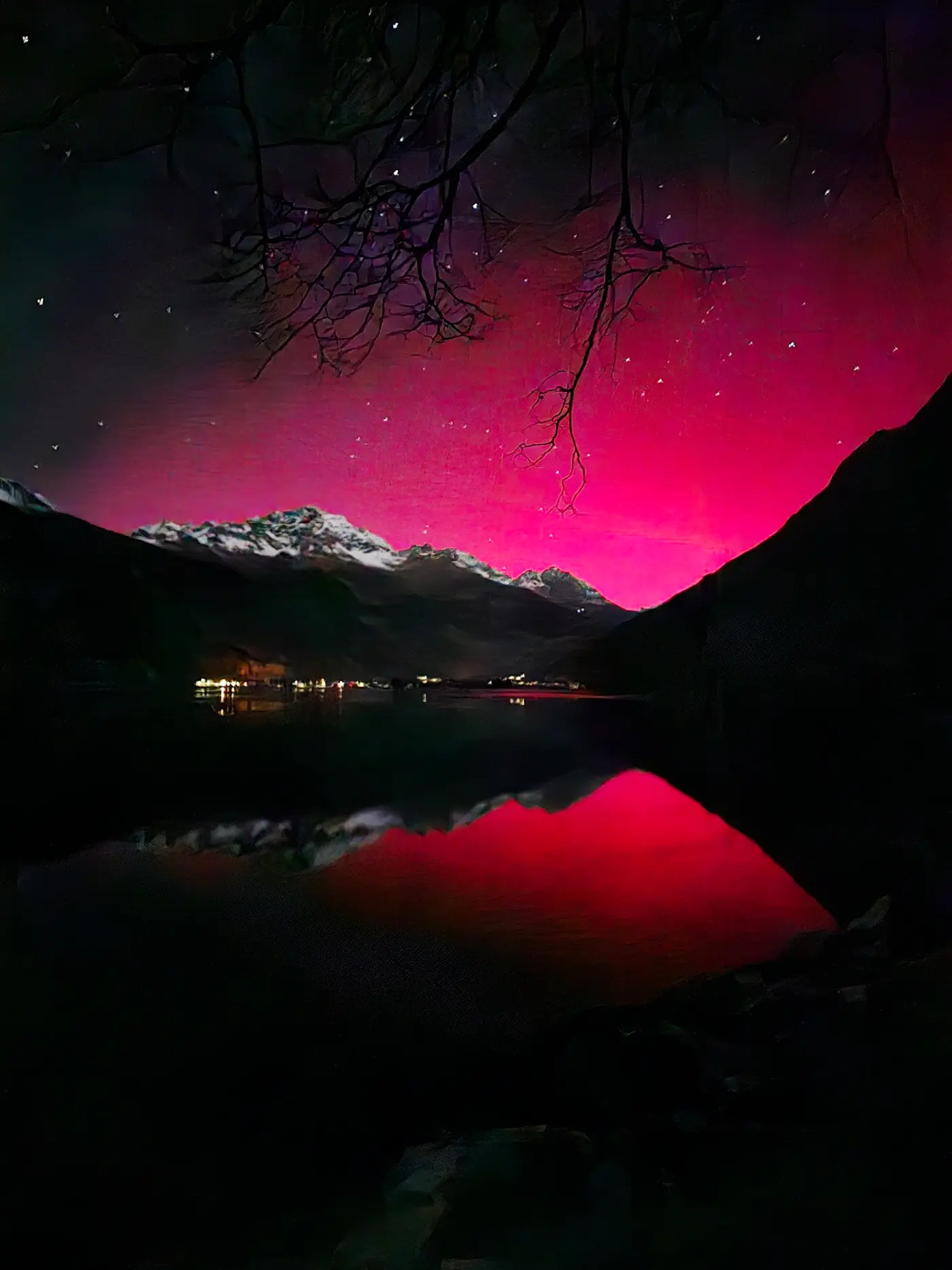 Un paesaggio notturno montano con un cielo rosa brillante, riflesso in un lago calmo. Le montagne innevate si stagliano all'orizzonte, mentre i rami di un albero si intravedono in alto. Stelle luminose punteggiano