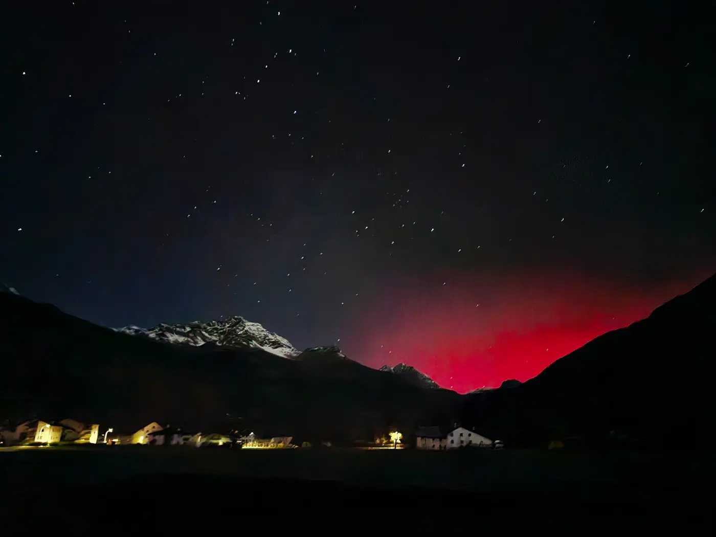 Cielo notturno stellato sopra un paesaggio montano, con un'illuminazione rossastra all'orizzonte e villaggi illuminati in basso.