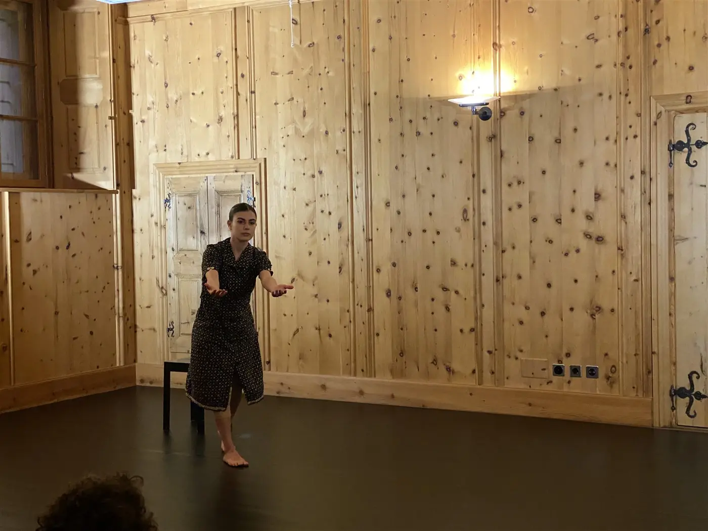 Una persona in abiti scuri si esibisce in una stanza con pareti in legno. È in piedi accanto a una sedia e gesticola con le mani. Il pavimento è scuro e c'è un'illuminazione soffusa.