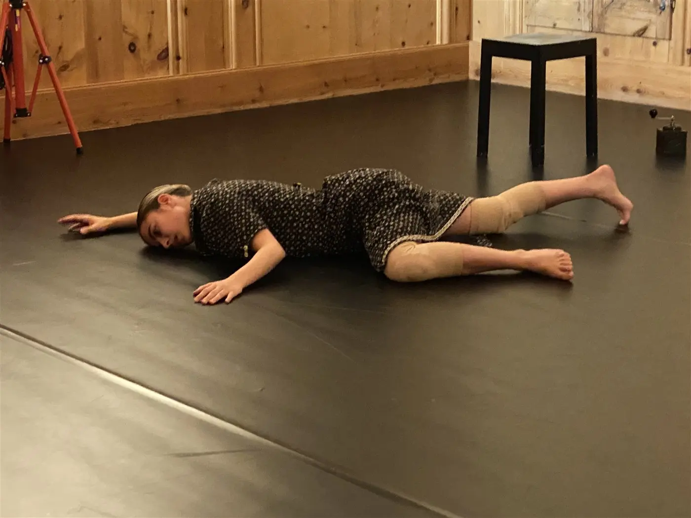 Una persona si trova distesa su un pavimento nero, in una posizione sdraiata, con le braccia estese e il volto rivolto verso il basso. Indossa un abito a pois e sembra concentrata in una performance o esercizio. Sullo sfondo ci sono elementi di