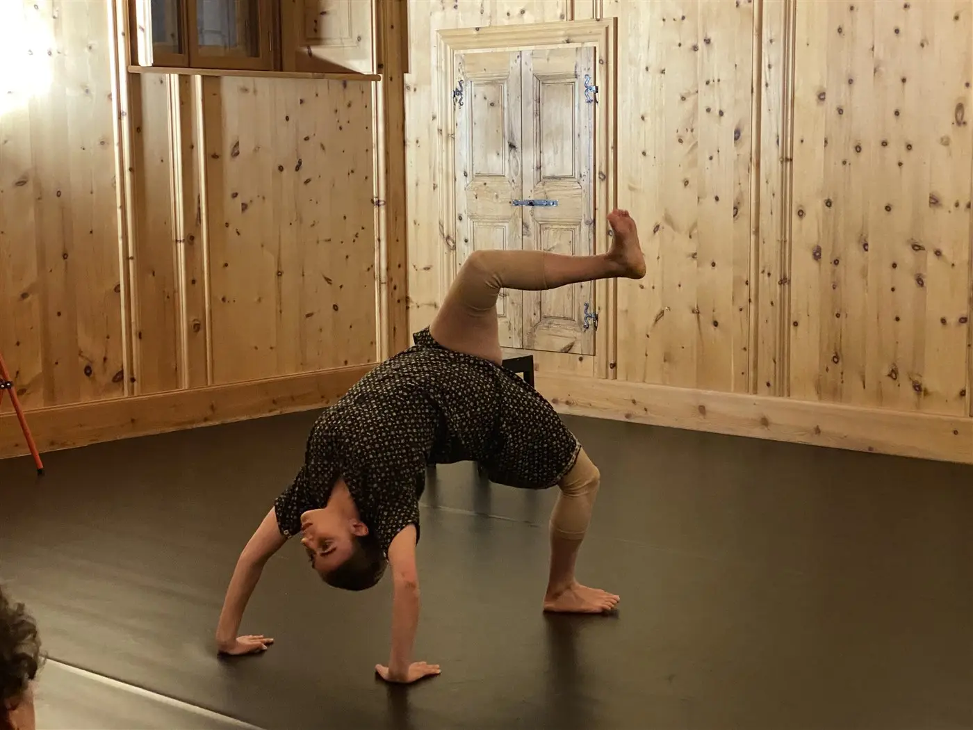 Un ballerino esegue una posa acrobatica su un pavimento nero in una stanza con pareti di legno. La figura è inclinata all'indietro, sostenendosi su una mano e una gamba sollevata.
