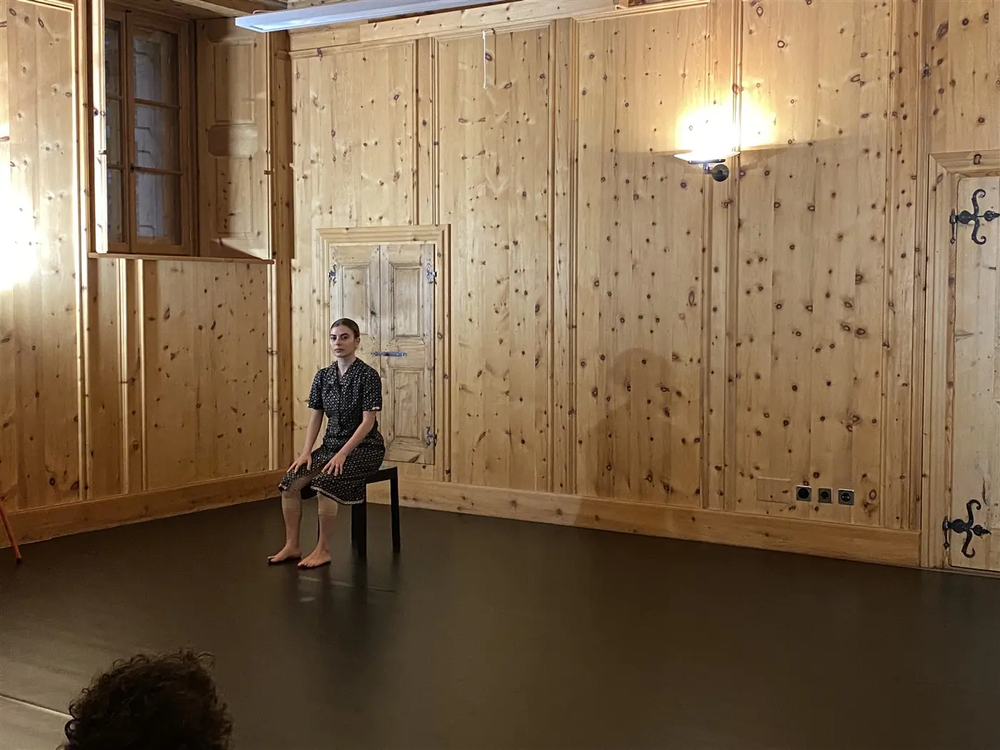 Una giovane donna seduta su una sedia in una stanza con pareti di legno. Indossa un vestito nero a pois bianchi. La stanza è vuota, con una luce soffusa e una finestra in alto a sinistra.