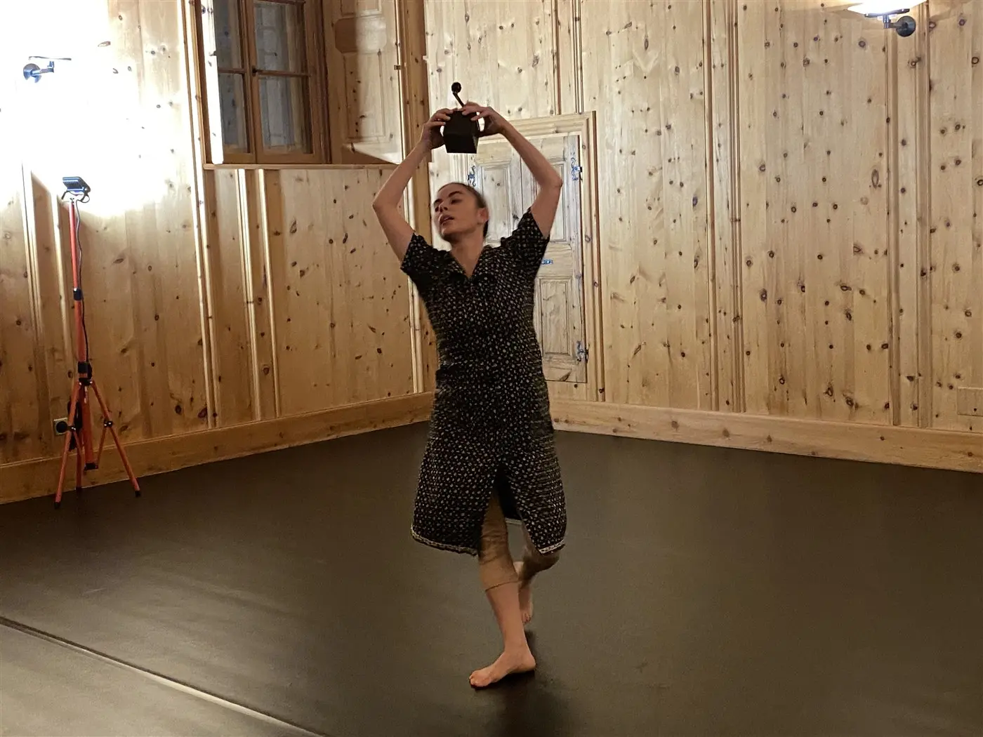 Una ballerina si esibisce in una stanza con pareti di legno, sollevando un oggetto sopra la testa mentre danza. La superficie è nera e ci sono luci di scena visibili.