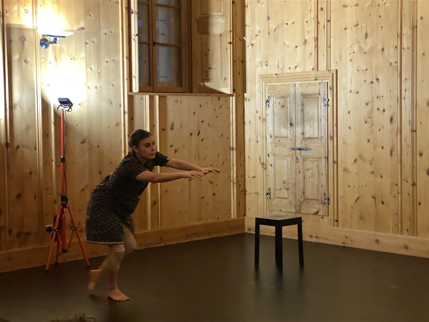 Una ballerina si esibisce in una sala con pareti di legno. Indossa un vestito a pois e si avvicina a uno sgabello nero con un gesto dinamico. La luce illumina l'ambiente e si intravedono finestre in legno.