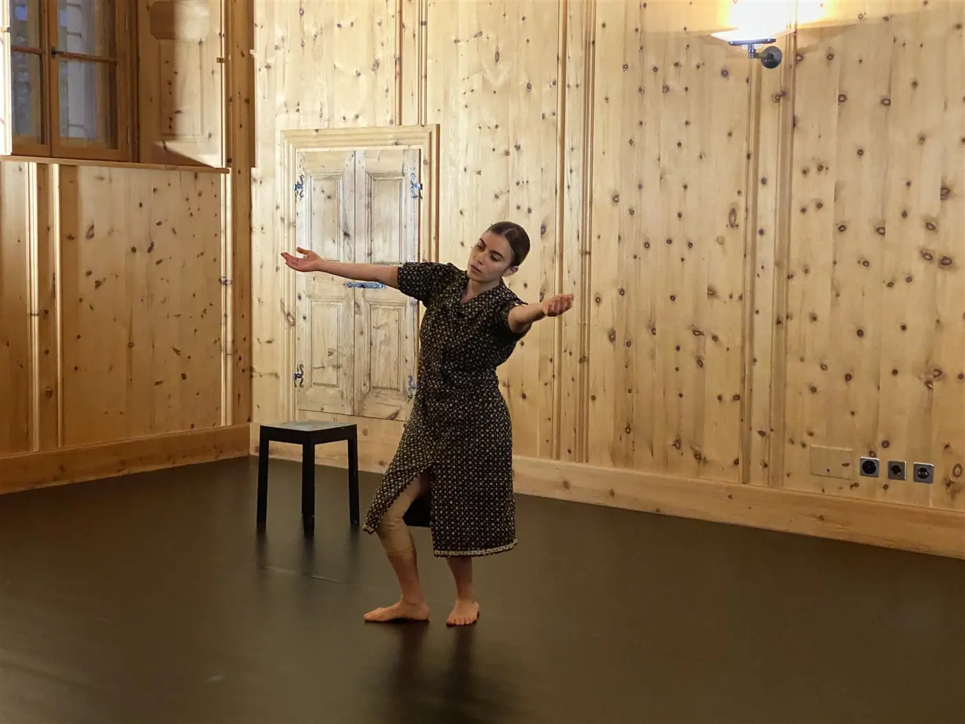 Una ballerina si esibisce in una stanza di legno, indossando un vestito a pois, con le braccia aperte e una gamba sollevata. Una sedia nera si trova accanto a lei.