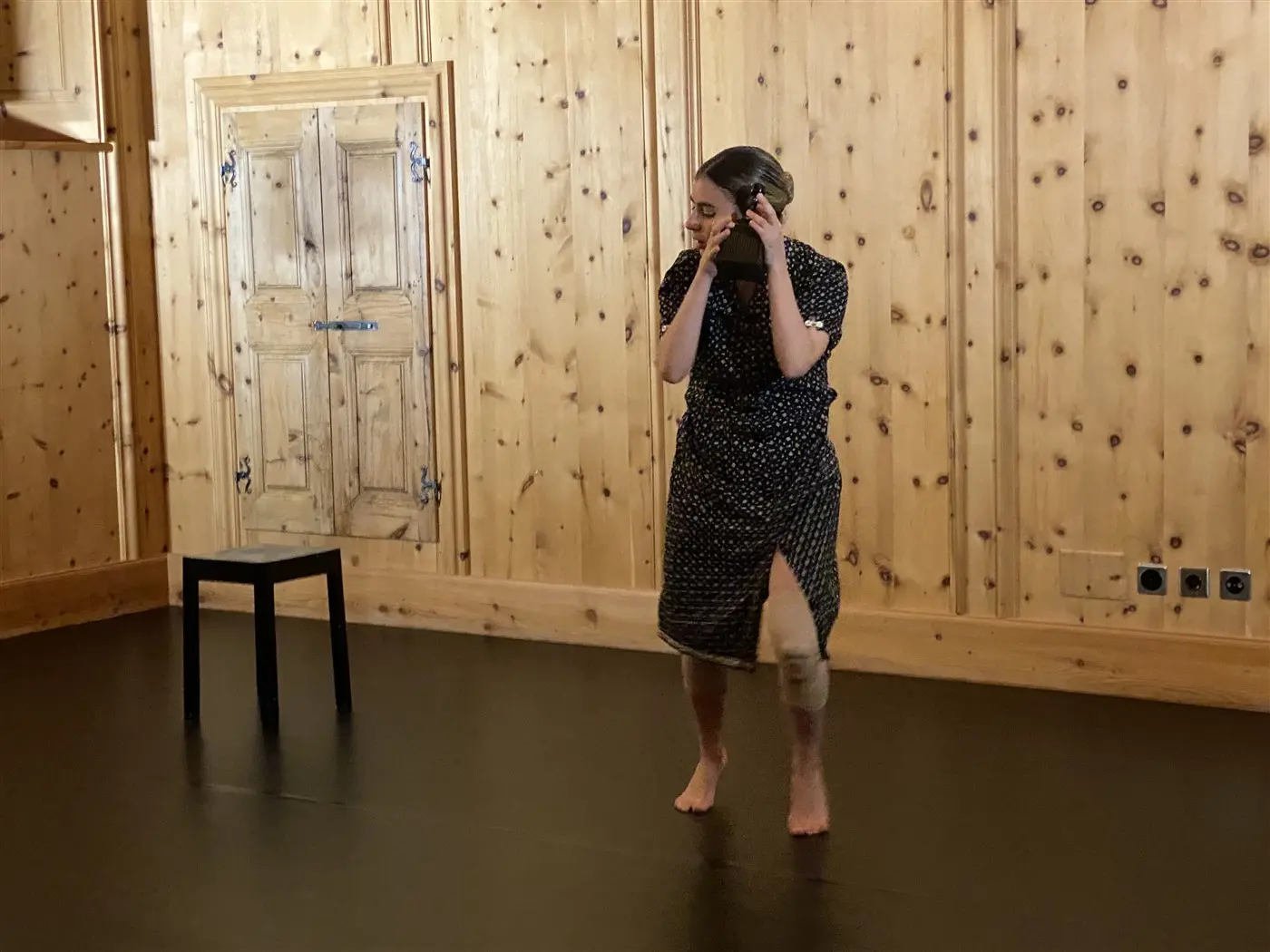 Una ballerina in abito a pois danza a piedi nudi in una stanza con pareti di legno. Accanto a lei c'è una sedia nera. L'atmosfera è intima e riflessiva.