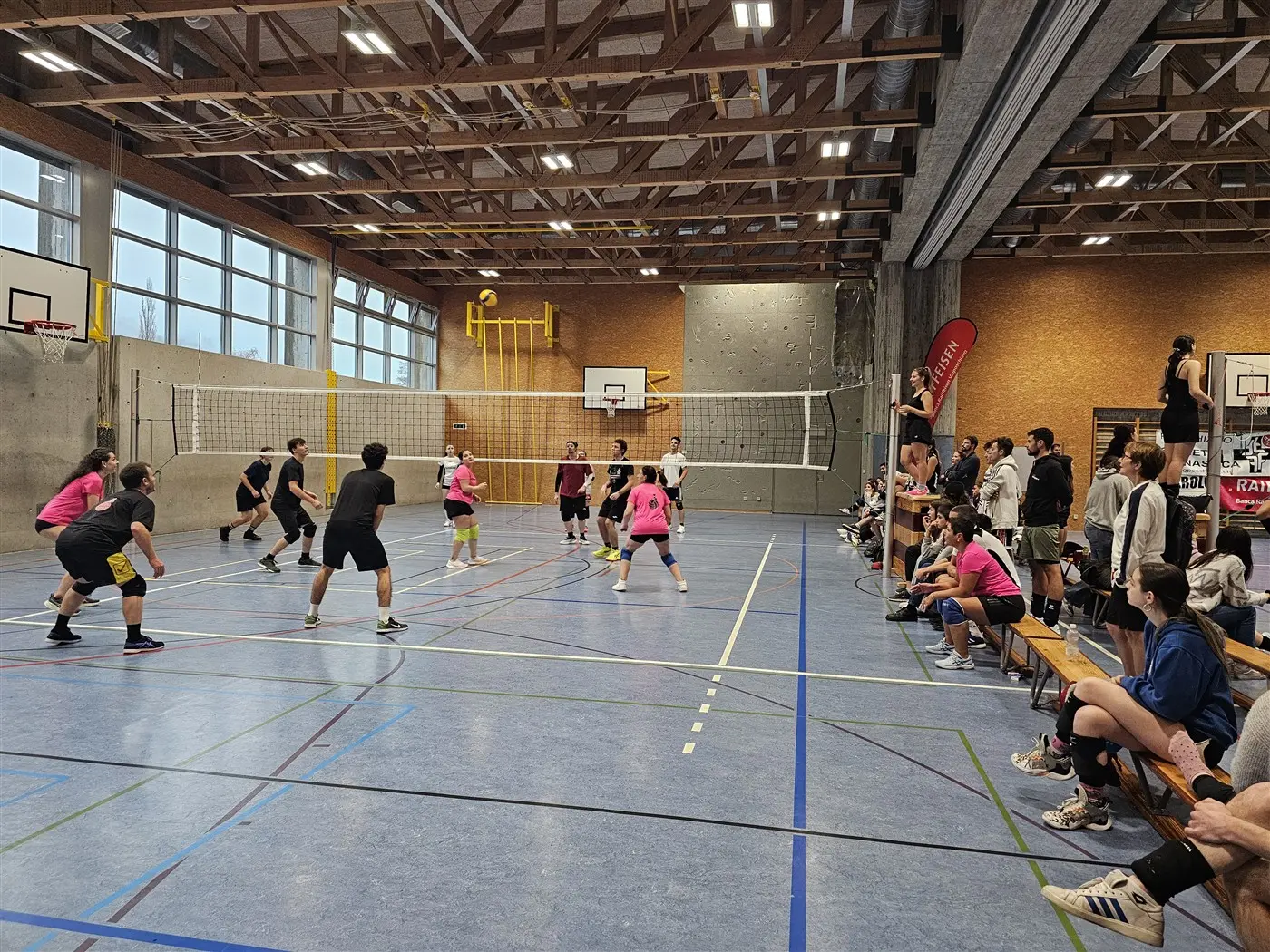 Una partita di pallavolo in un palazzetto. Giocatori in magliette rosa e nere sono in azione, mentre a bordo campo ci sono spettatori che seguono il gioco. Il campo è attrezzato con reti e canestri.