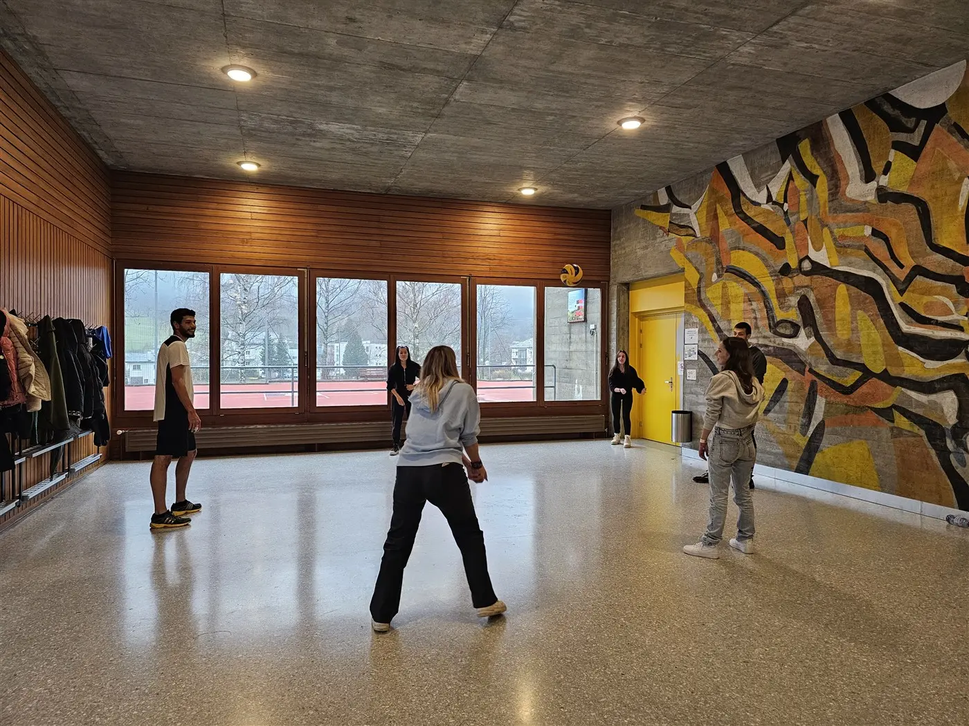 Un gruppo di sei persone si allena in un'area interna spaziosa, con pareti di legno e un grande murale colorato. Alcuni partecipanti sembrano impegnati in un'attività di riscaldamento. Fuori dalle finestre si intravede un pa