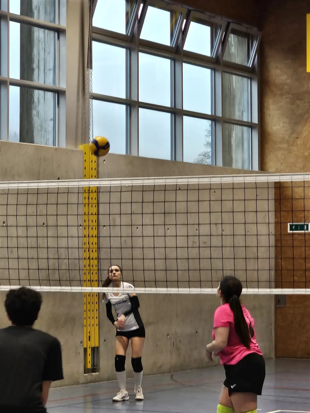 Due giocatrici di volley si preparano a ricevere la palla mentre un compagno serve. Si intravede una rete alta tra di loro e una parete con finestre sullo sfondo.