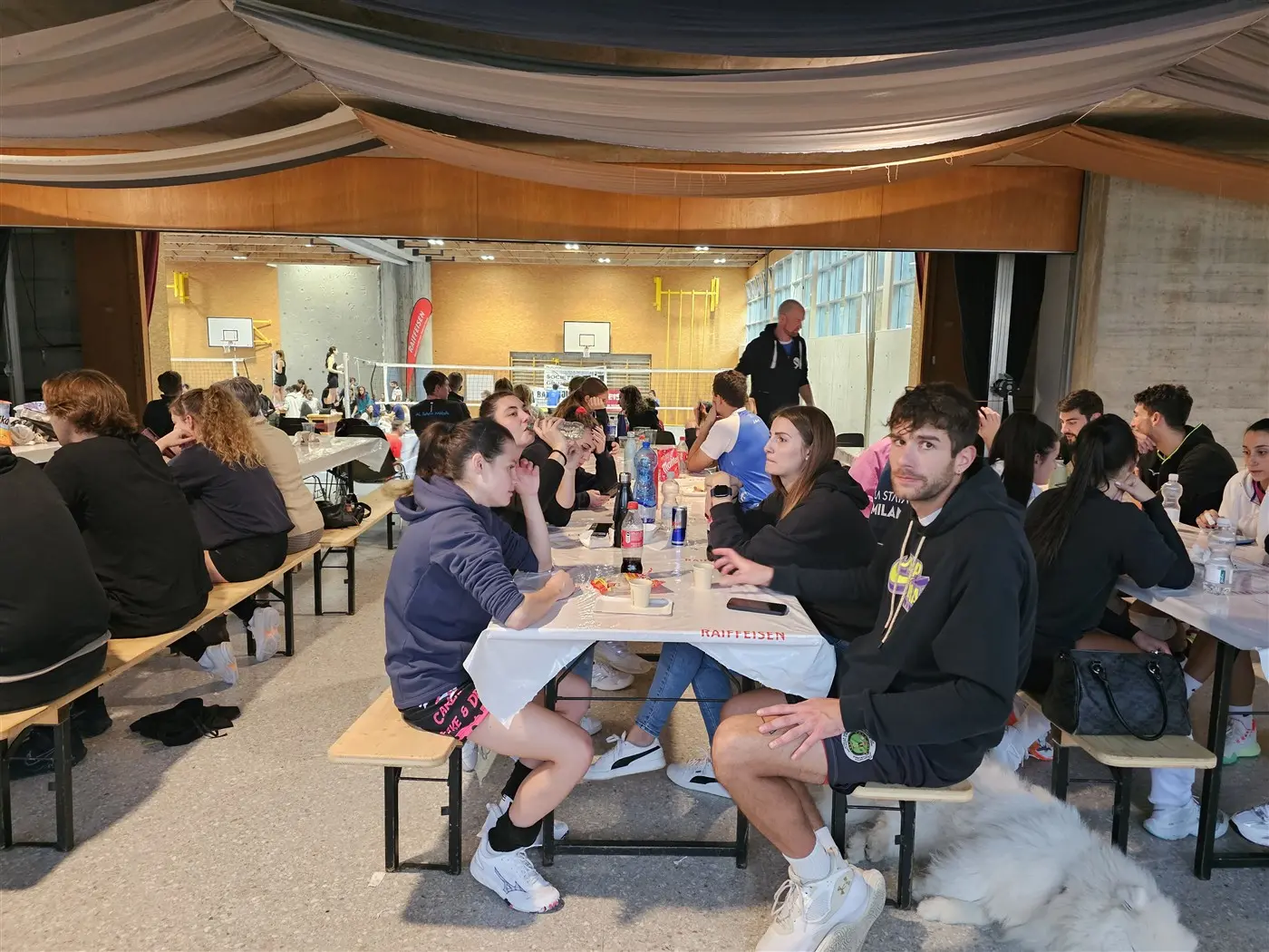 Gruppo di giovani seduti a un tavolo in una sala, intenti a conversare e mangiare. In fondo si vedono campi da gioco e altri partecipanti che si divertono. Arredamento semplice con decorazioni appese.