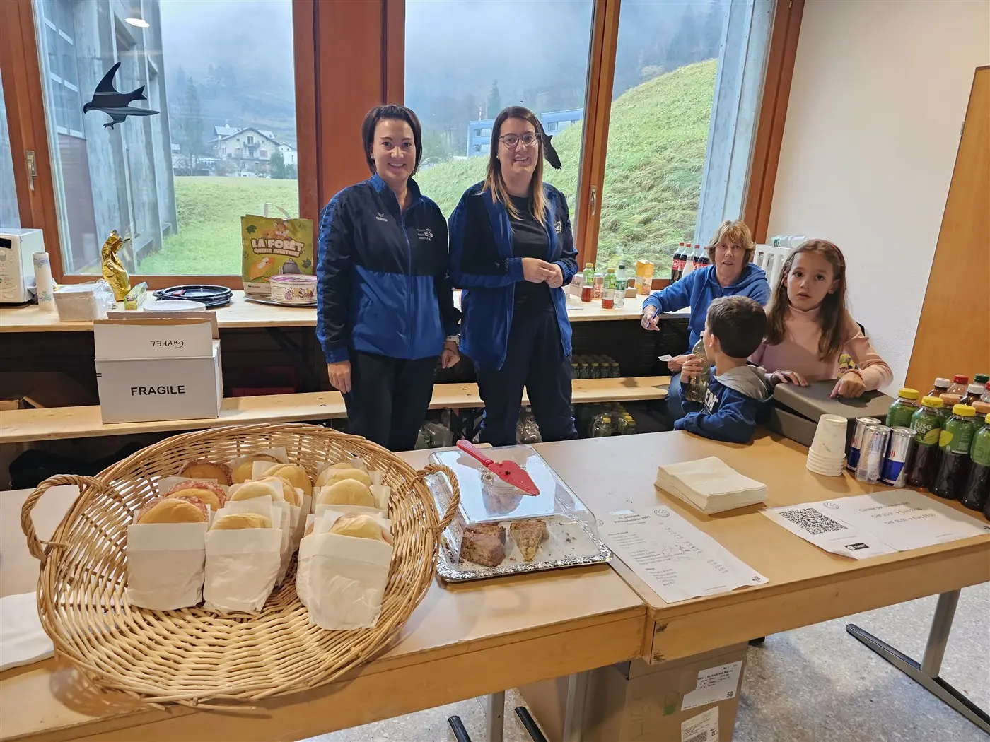 Due donne sorridenti si trovano dietro un banco con un cestino di panini e diversi cibi e bevande. Sullo sfondo, si intravedono bambini e un paesaggio verde attraverso le finestre.