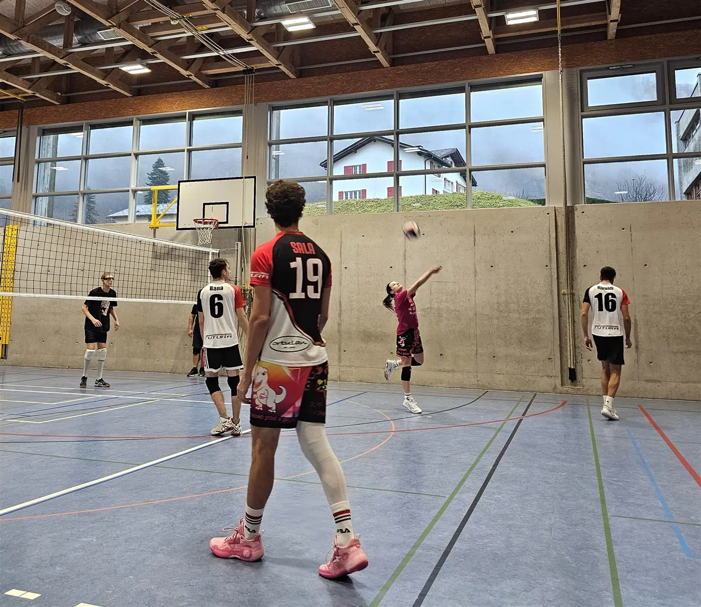 Giocatori di pallavolo in un palazzetto. Un giocatore sta per servire, mentre gli altri sono pronti a ricevere il colpo. Le maglie dei giocatori sono diverse, con numeri visibili. Dietro si vedono finestre ampie con una
