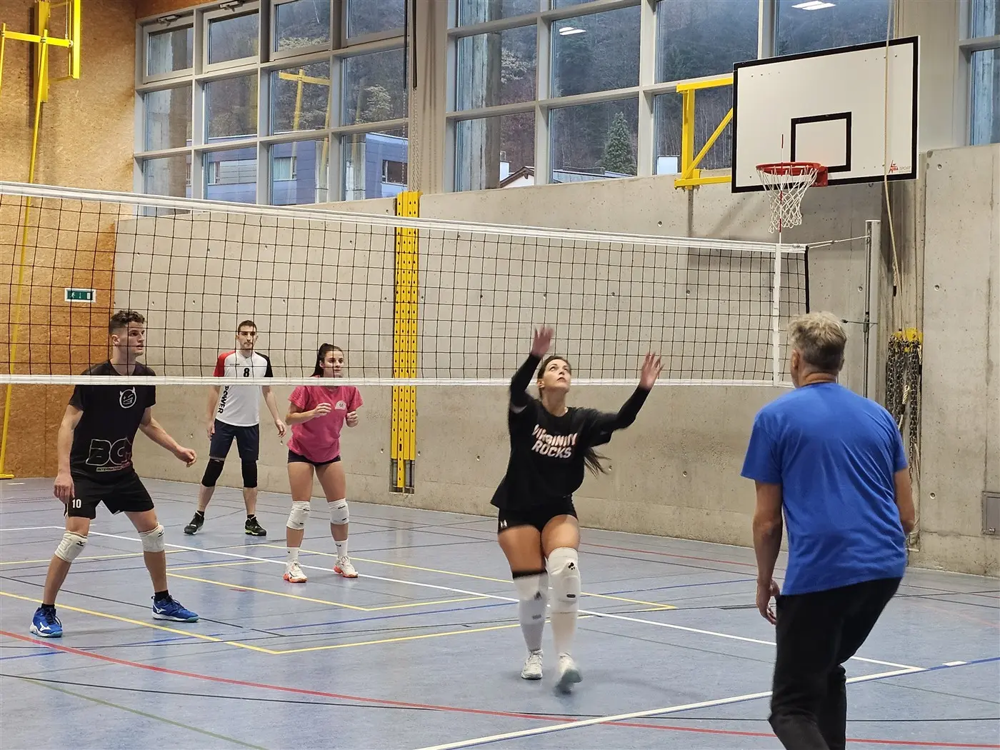 Un gruppo di quattro giocatori di pallavolo in campo. Una giocatrice si prepara a ricevere la palla, mentre gli altri osservano. Si notano reti e linee di gioco, con un ambiente interno ben illuminato.