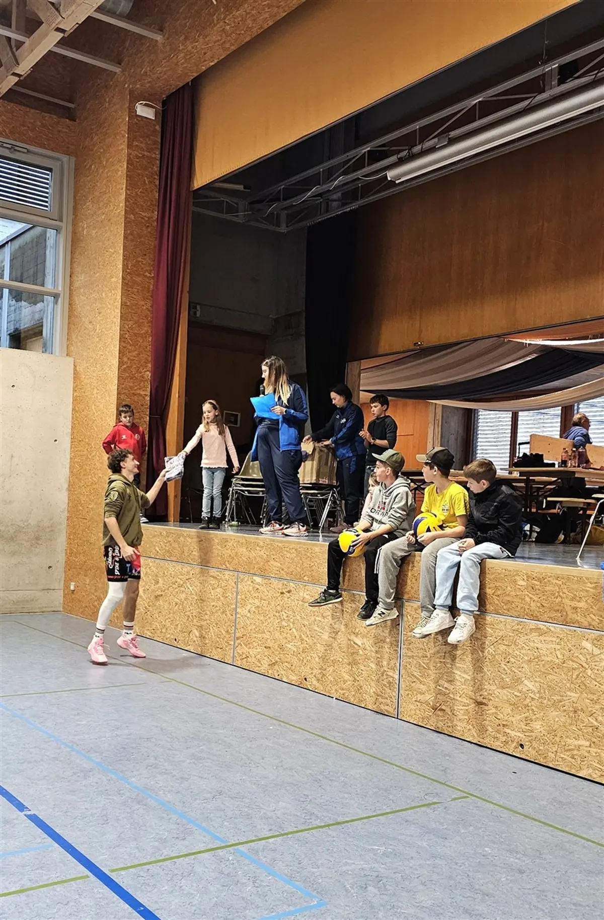 Un gruppo di bambini e ragazzi si trova in una palestra. Alcuni sono seduti su un rialzo, mentre altri stanno in piedi. Una ragazza sta passando un pallone a un’amica in divisa sportiva. L'ambiente è luminoso, con pareti in legno e