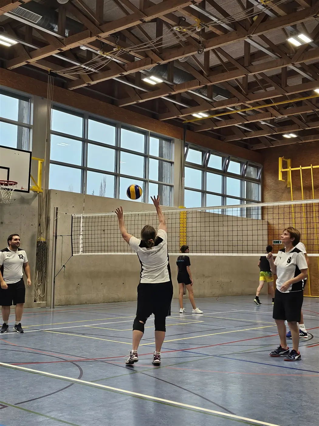 Un gruppo di persone sta giocando a pallavolo in una palestra. Una giocatrice si prepara a fare un palleggio mentre altre due si avvicinano e un’altra si trova in fondo. Le pareti sono di cemento e ci sono ampie finestre che lasciano entra