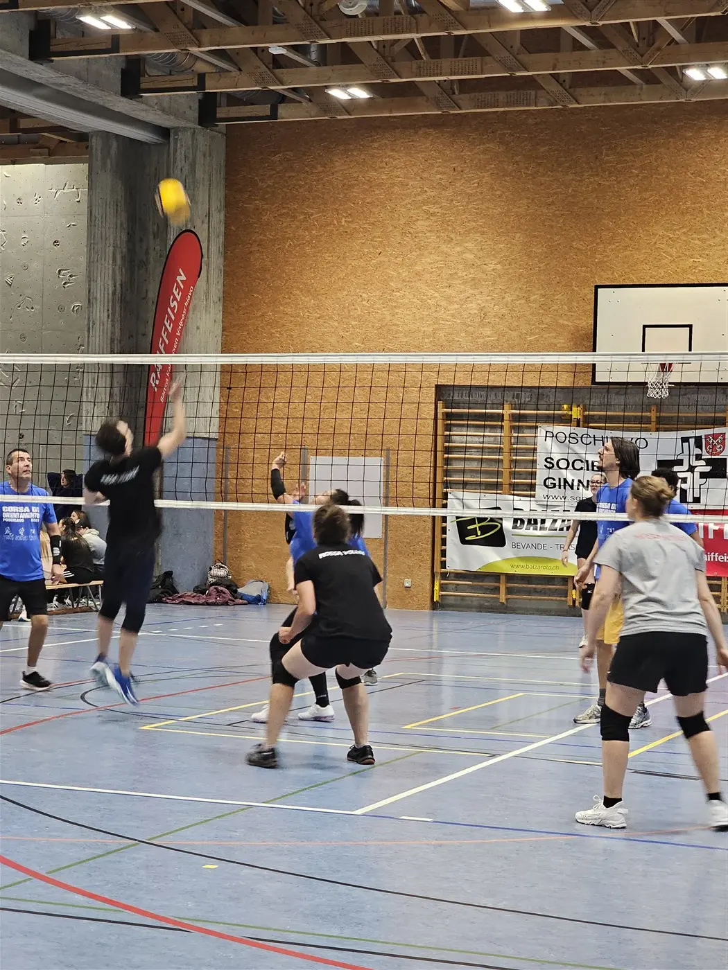 Un momento di gioco in una partita di pallavolo indoor. Un giocatore in maglietta blu salta per colpire la palla, mentre gli altri compagni di squadra si preparano in campo. Sullo sfondo, ci sono banner e una rete da pallavolo