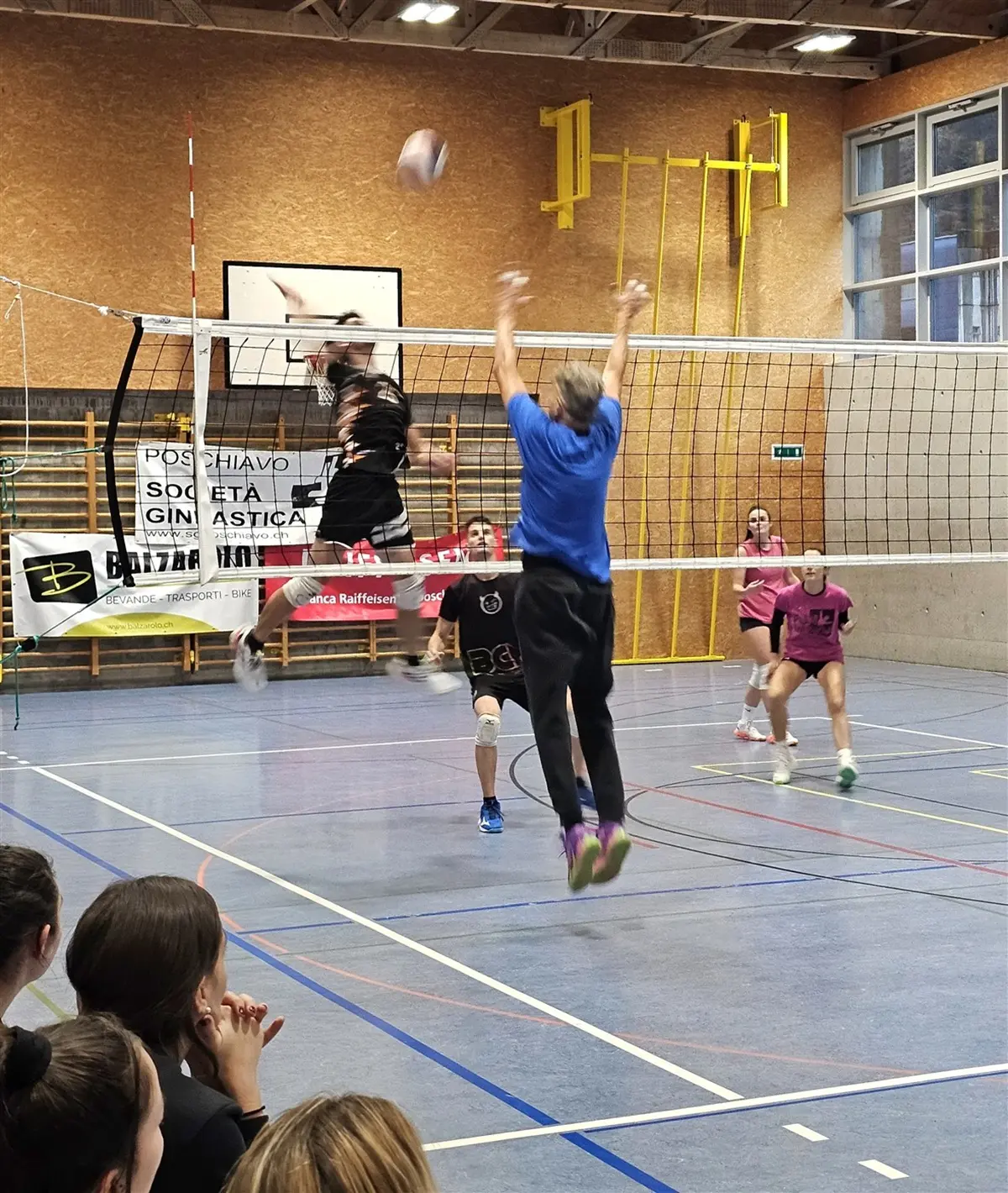 Giocatori di pallavolo in un'azione di gioco. Un atleta salta per schiacciare mentre un avversario tenta di bloccare il colpo. Alcuni compagni di squadra osservano dalla panchina.