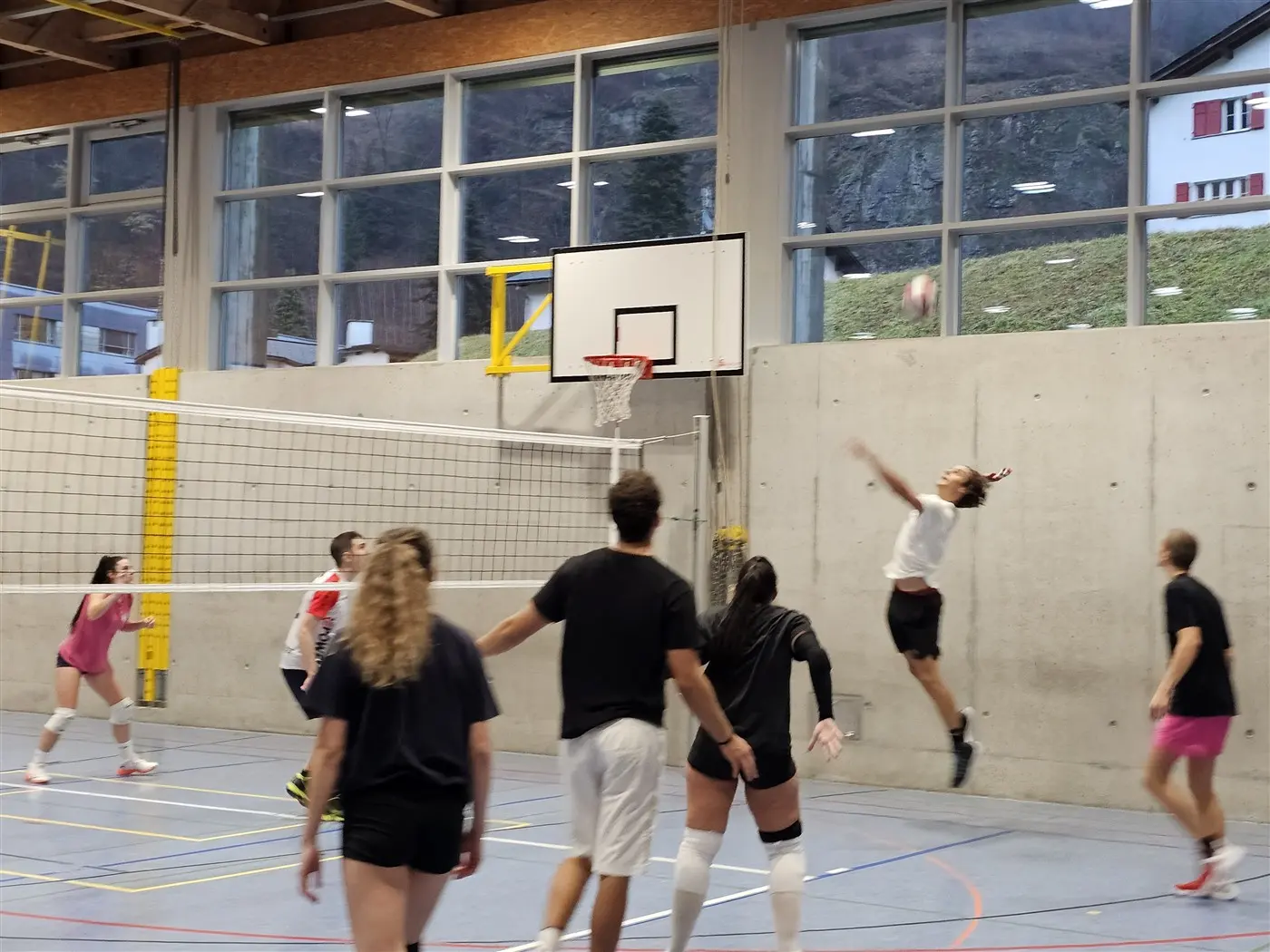 Giocatori di pallavolo in palestra durante un allenamento. Una giocatrice salta per colpire la palla sopra la rete, mentre altre sono in attesa in diverse posizioni. Il campo è illuminato da grandi finestre.