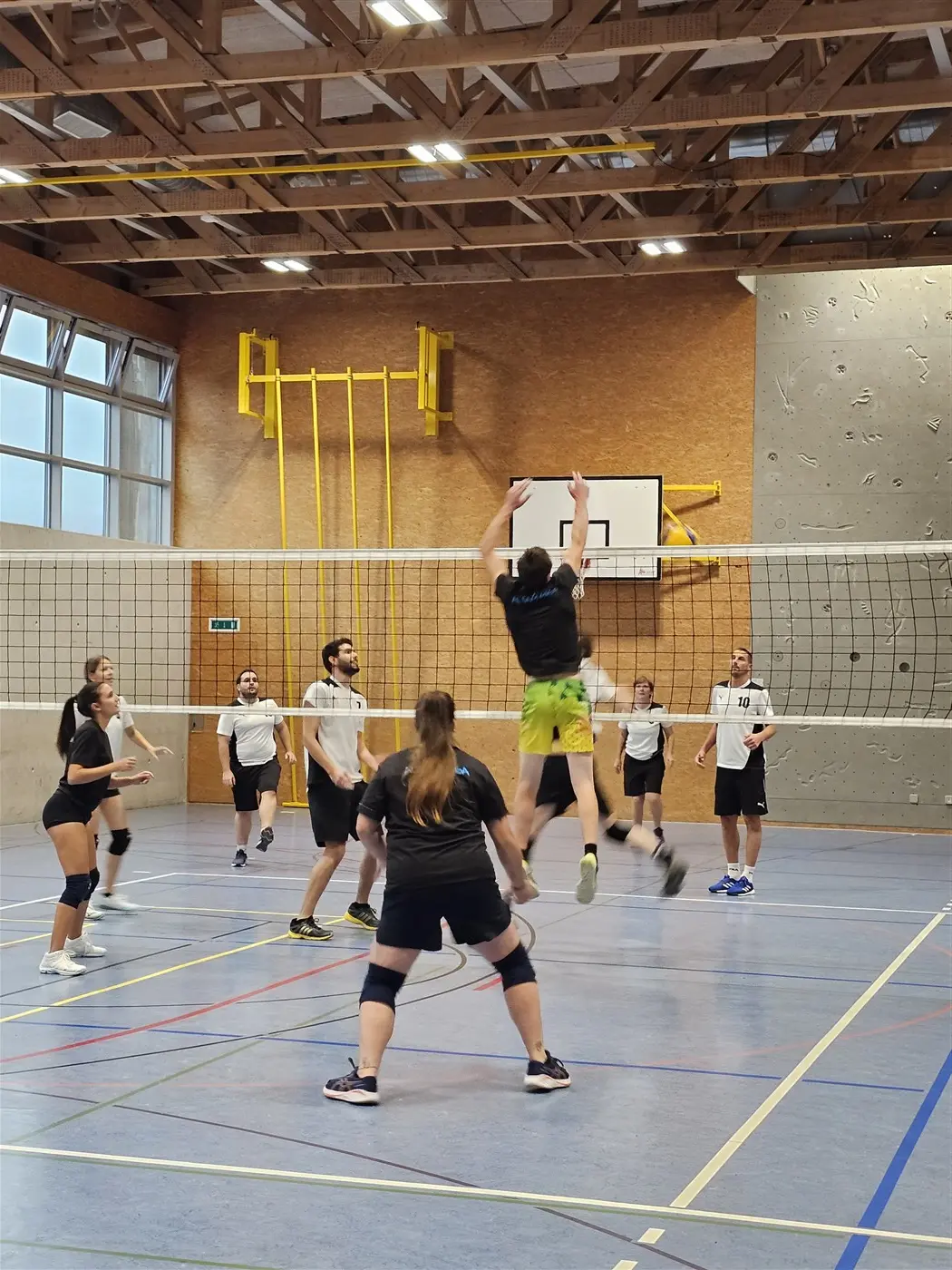 Un gruppo di persone gioca a pallavolo in un ambiente interna, con una rete al centro. Un giocatore salta per colpire la palla durante un'azione di gioco, mentre gli altri sono in attesa e pronti a ricevere.
