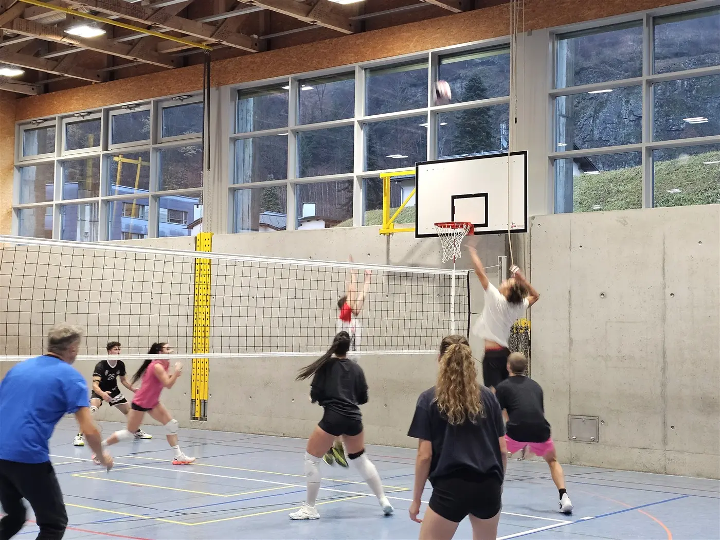 Giocatori di pallavolo in azione in un campo al coperto. Una persona salta per colpire la palla sopra la rete, mentre gli altri si preparano per la ricezione. Gran parte della sala è illuminata da grandi finestre.
