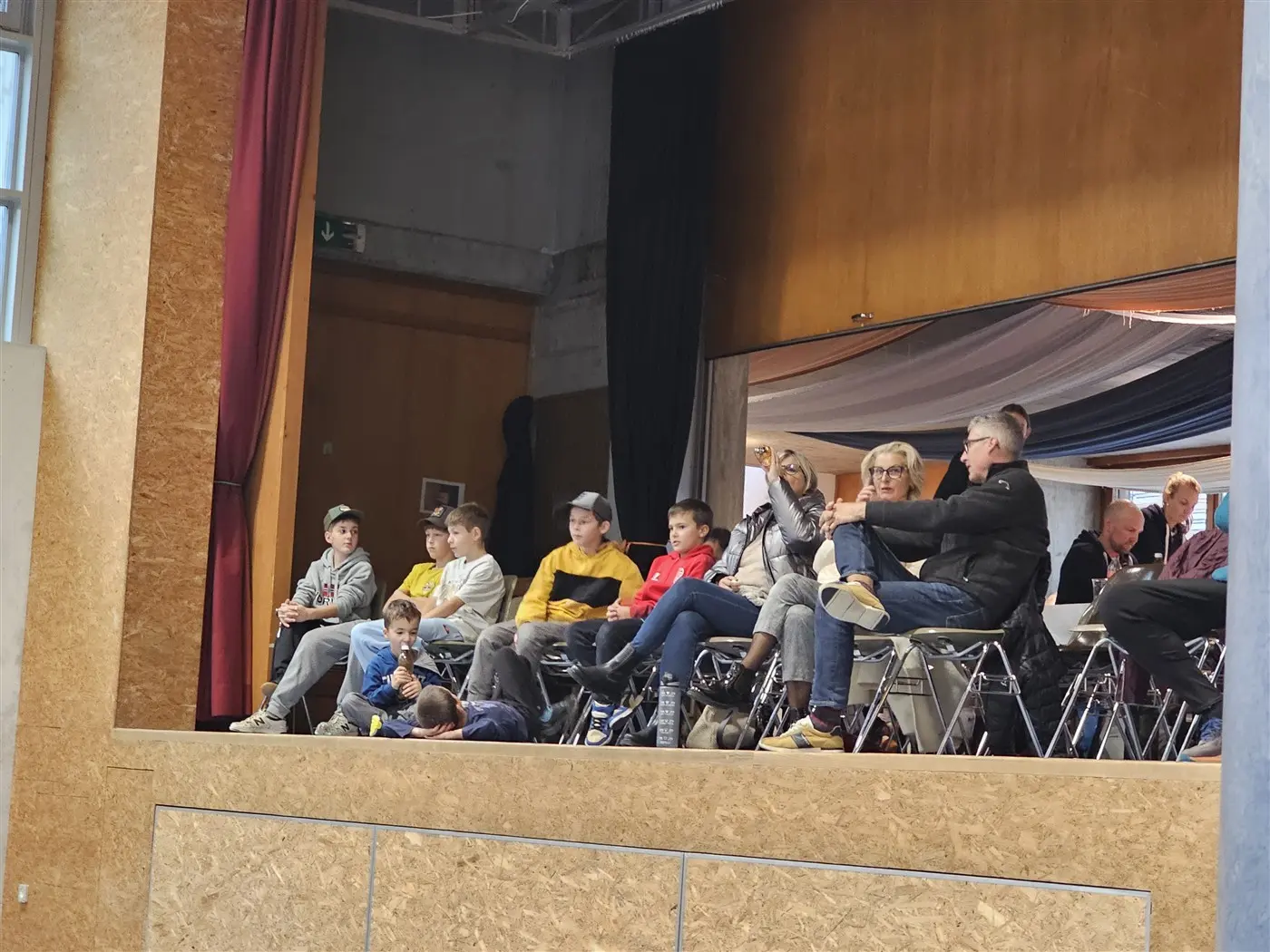 Un gruppo di persone sedute su una piattaforma, tra cui bambini e adulti, che osservano un evento in sala. La scena è caratterizzata da un ambiente informale e da un'atmosfera conviviale.