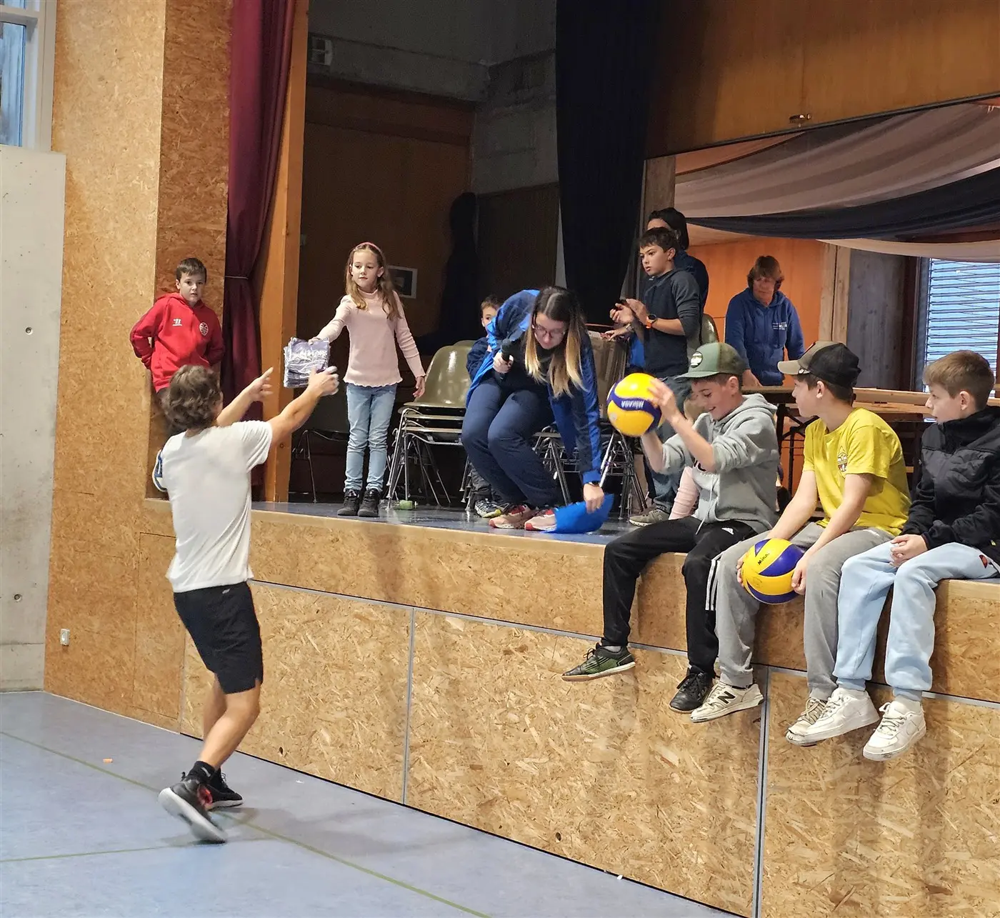 Un ragazzo lancia una palla da volley mentre un gruppo di bambini e ragazzi osserva dalla parte alta della sala. Alcuni sono seduti, mentre altri stanno in piedi.
