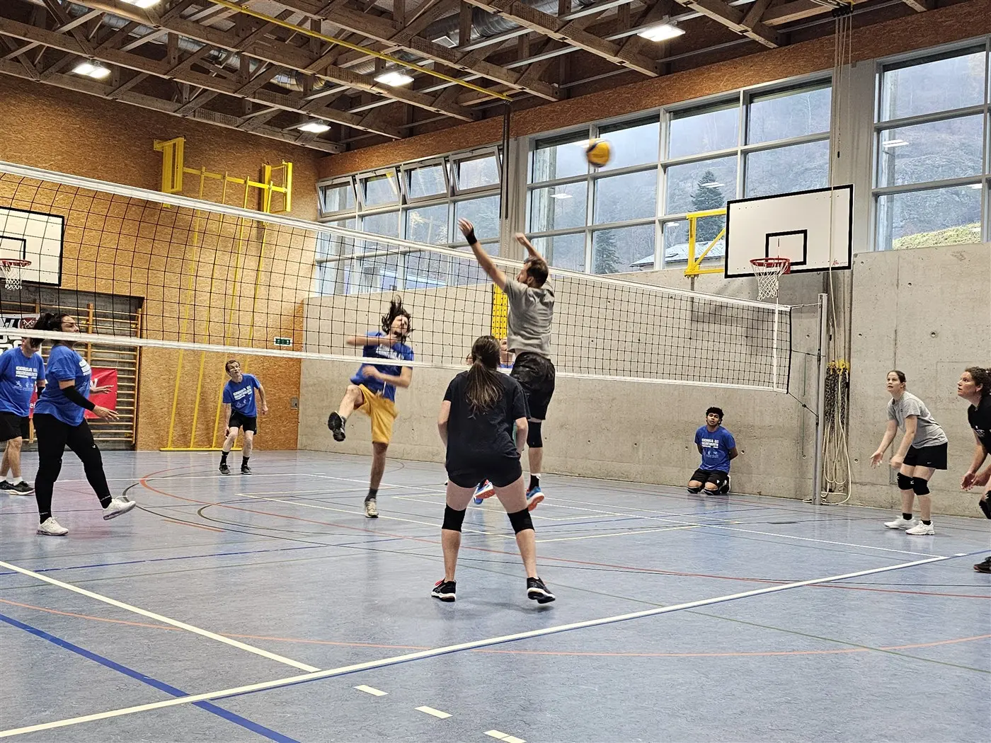 Giocatori di pallavolo in azione in una palestra. Una persona sta saltando per colpire la palla sopra la rete, mentre altri partecipanti sono in attesa o si preparano a ricevere. Il campo è ben illuminato.