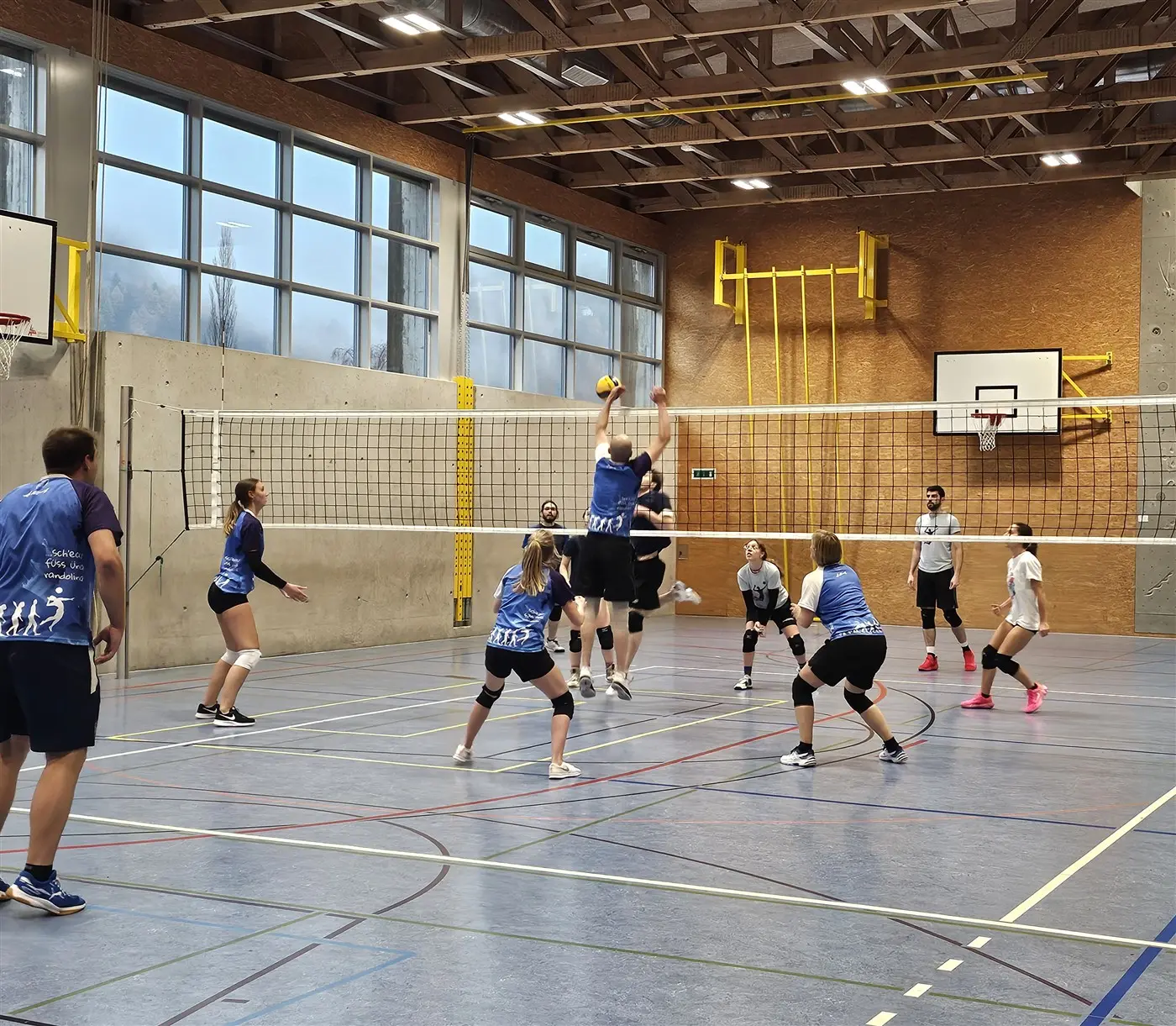 Una squadra di volley si allena in una palestra. Giocatori e giocatrici indossano divise blu e bianche. Un atleta sta saltando per colpire la palla mentre gli altri osservano. La rete e le linee del campo sono visibili in un