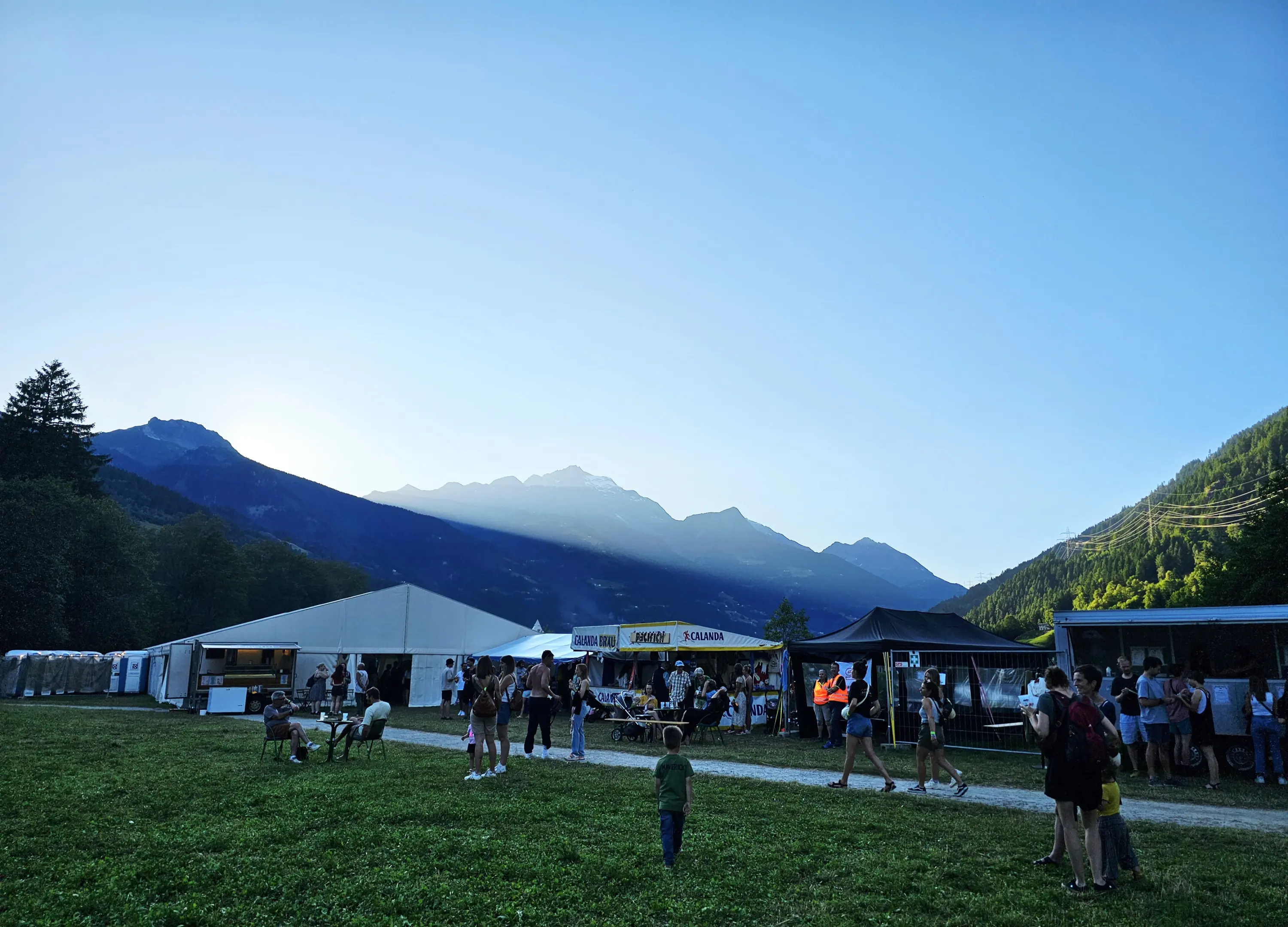Festa all'aperto con tende e persone in un prato, circondata da montagne illuminate dal sole al tramonto.