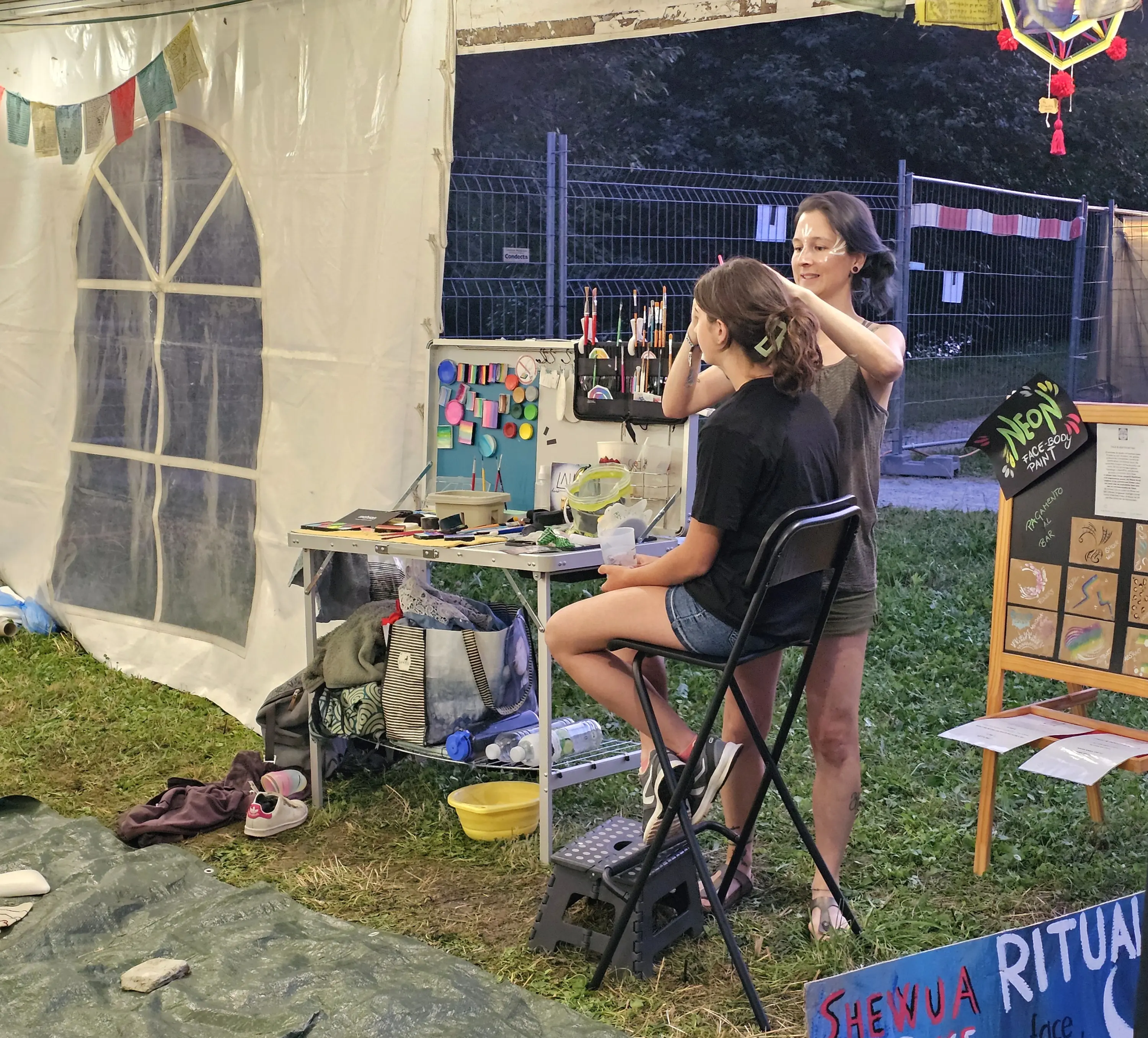 Due donne sono all'interno di una tenda. Una di loro sta sistemando i capelli dell'altra, seduta su uno sgabello. Sul tavolo ci sono vari strumenti e colori. Sullo sfondo si vedono tende e decorazioni.