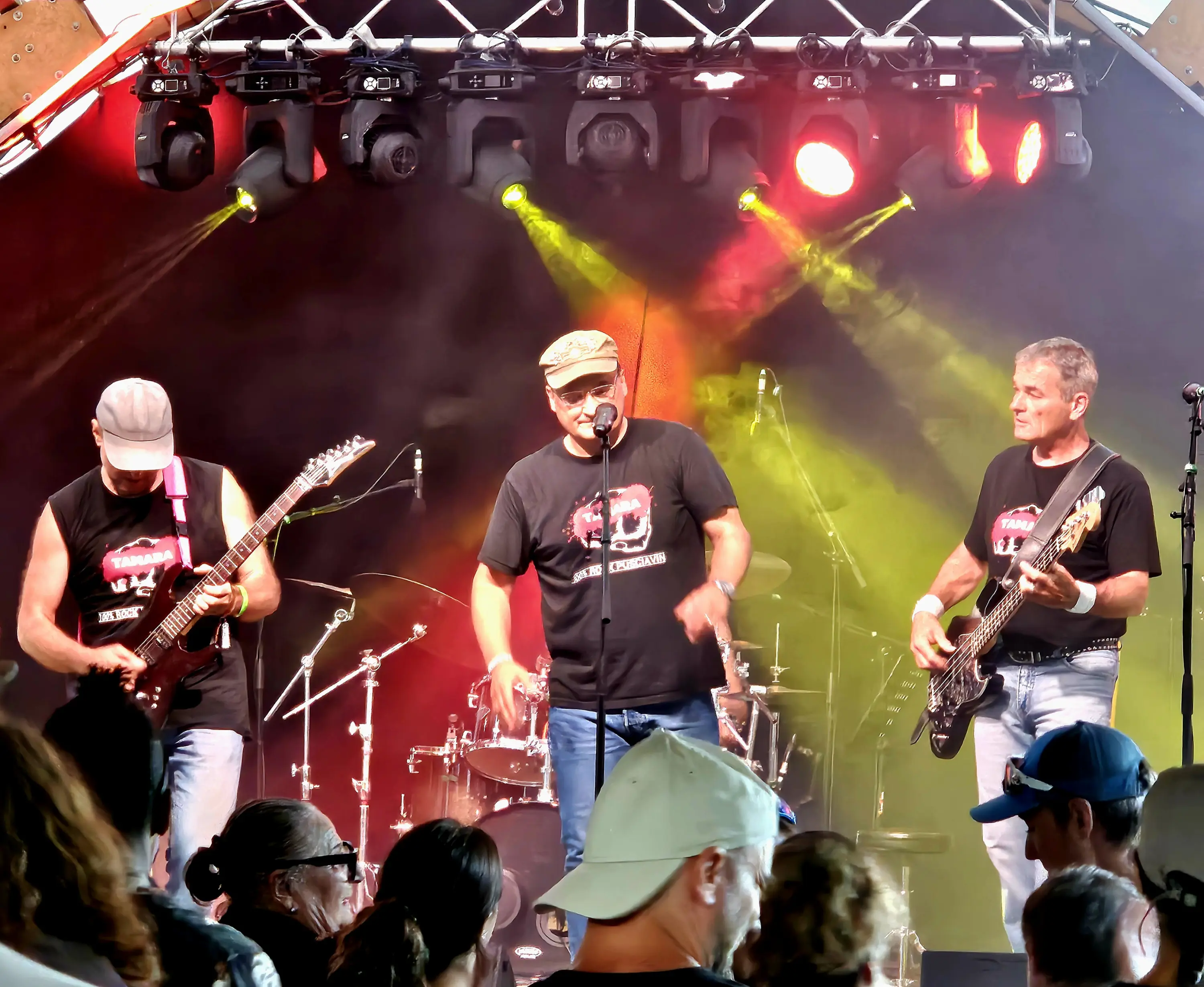 Tre musicisti si esibiscono su un palco. Uno suona la chitarra, il secondo ha un microfono e il terzo suona il basso. Sullo sfondo, una batteria e luci colorate illuminano l'atmosfera. Pubblico visibile in primo piano.