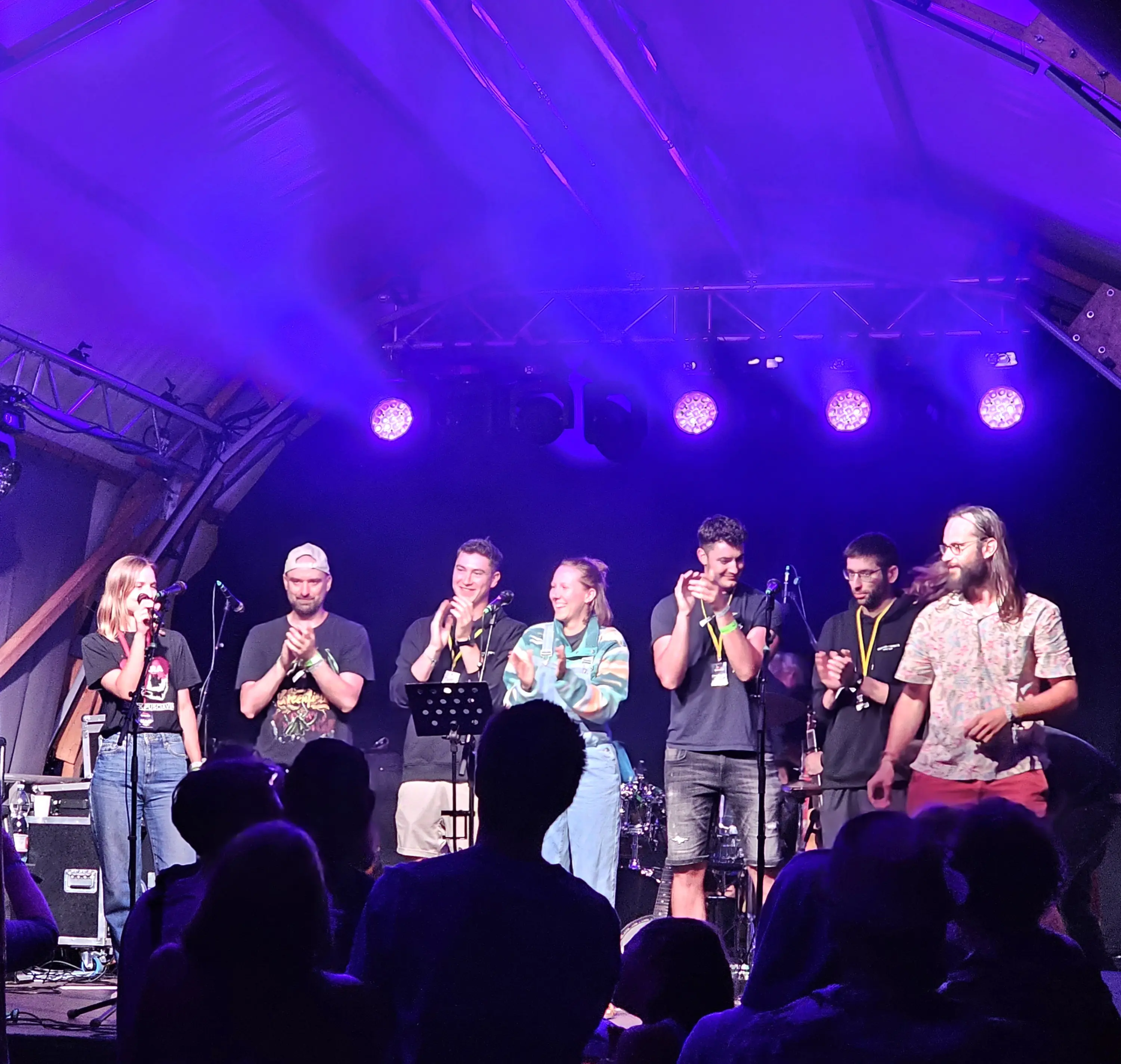 Un gruppo di sei giovani sul palco applaude, illuminati da luci colorate. Alcuni suonano strumenti musicali, mentre il pubblico inizia a rispondere con entusiasmo.