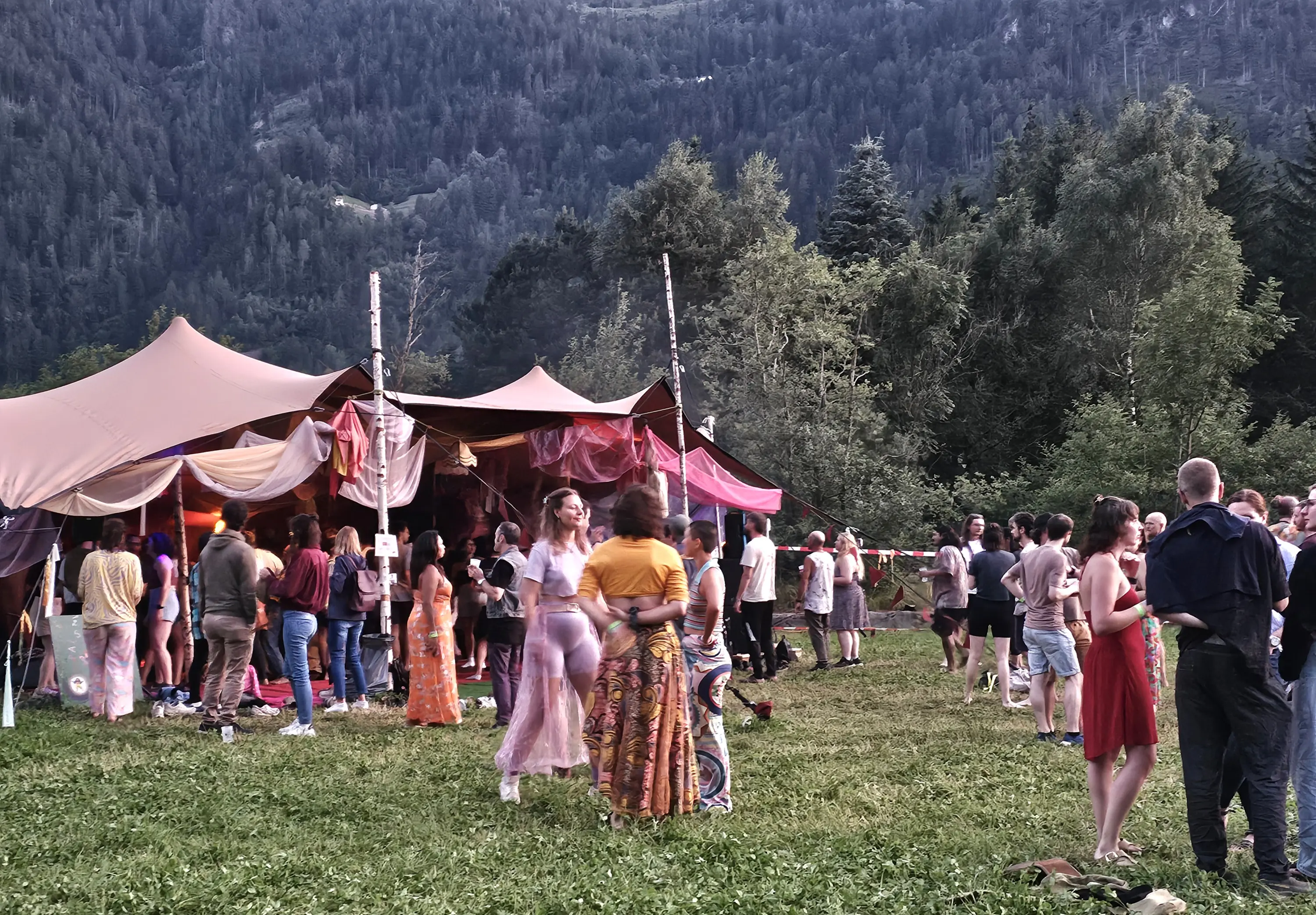 Un festival all'aperto con persone che ballano e socializzano. Si vedono tende colorate e un paesaggio montano sullo sfondo. Alcuni partecipanti indossano abiti estivi e ci sono gruppi di amici che si divertono.