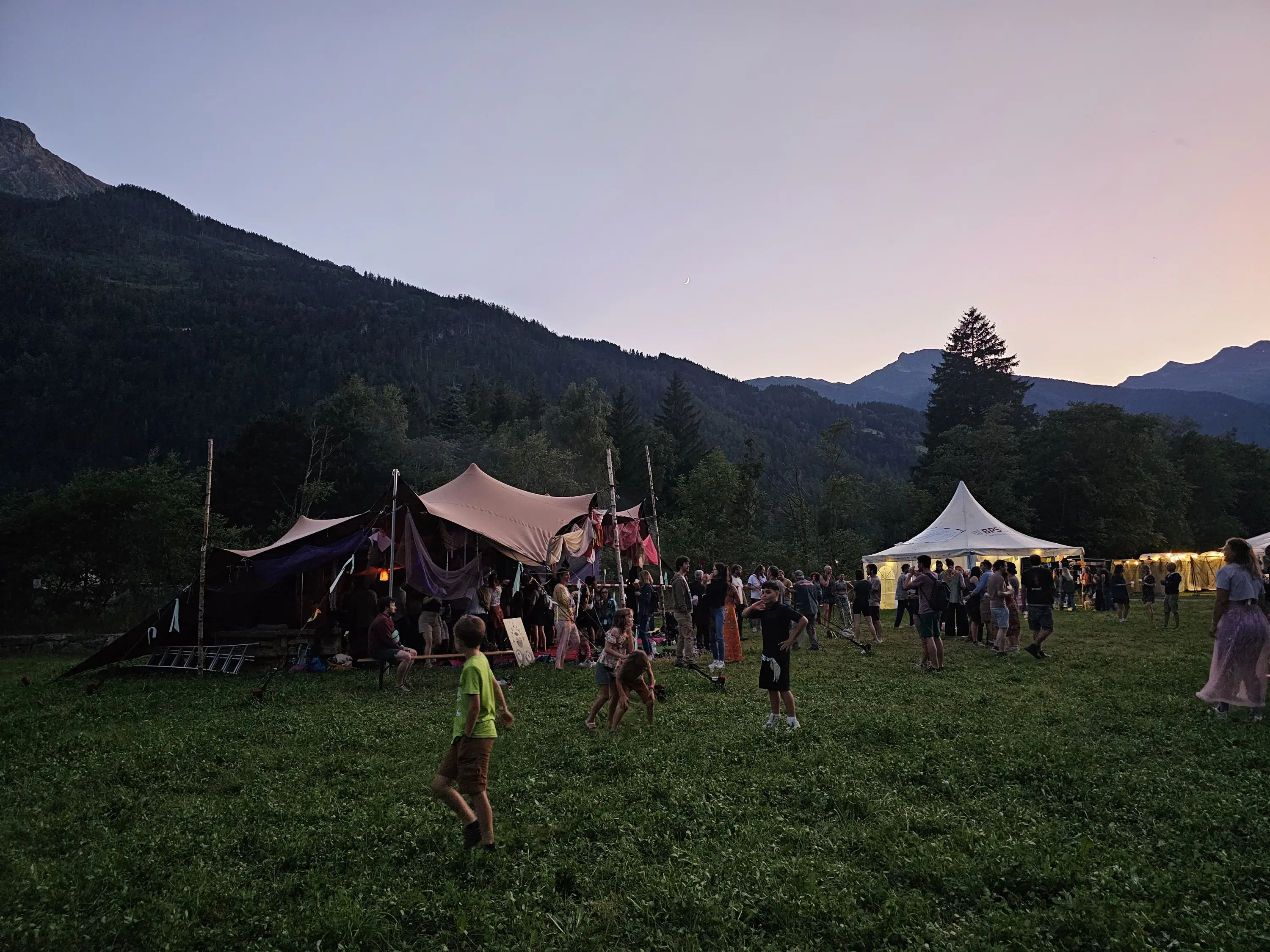 Un festival all'aperto al tramonto, con tende colorate e una grande tenda bianca. Gruppi di persone si divertono in un prato verde, circondati da montagne. Bambini giocano mentre gli adulti socializzano e si godono l'atmosfera.