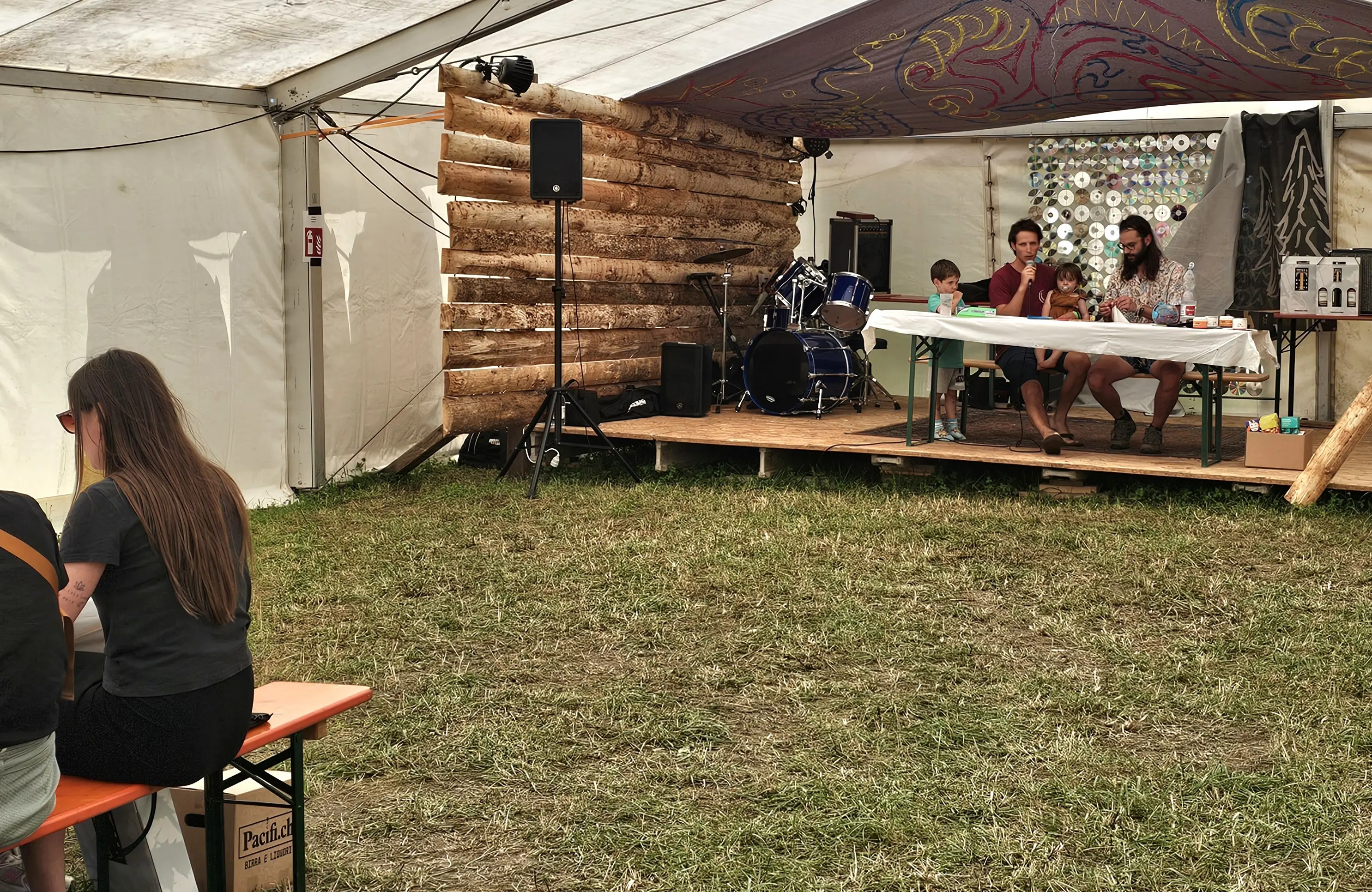 Un'area all'interno di una tenda con un palco in legno e strumenti musicali. Alcune persone sono sedute a un tavolo mentre altre sono in piedi. Sullo sfondo, decorazioni artistiche e un sistema audio. Il pavimento è di erba.