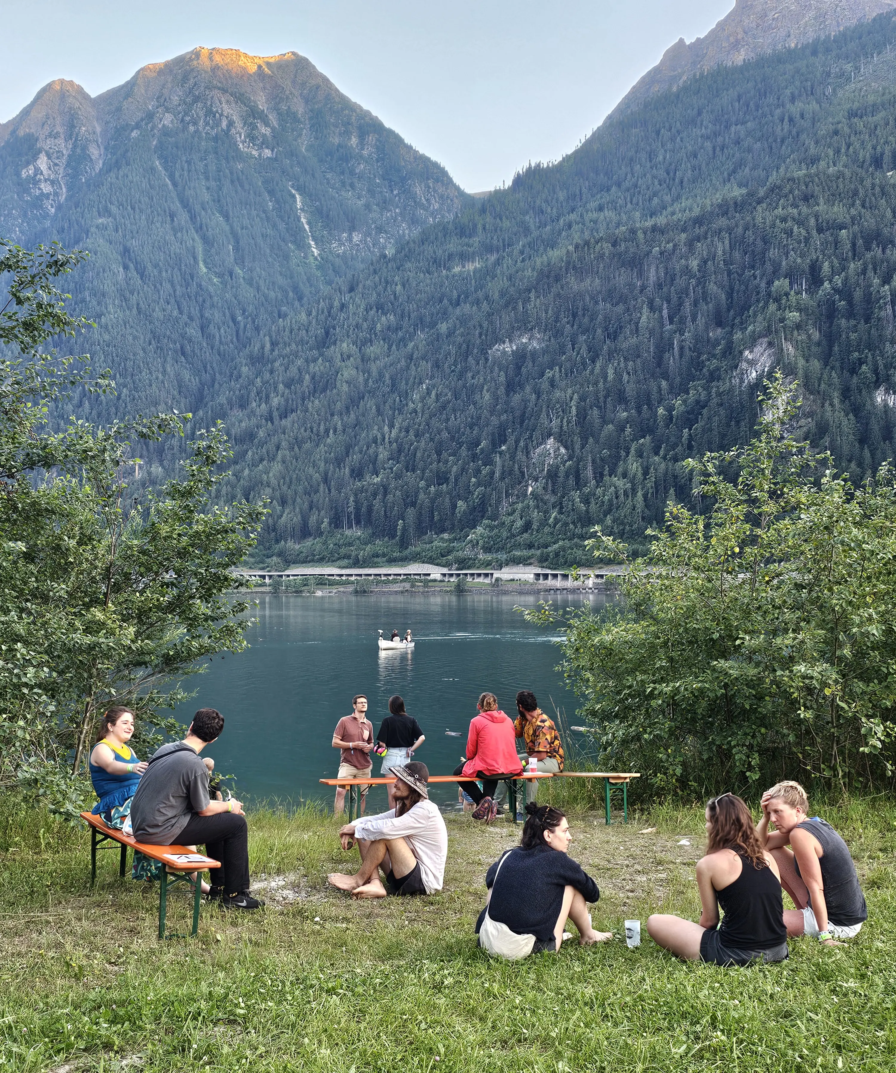 Un gruppo di persone si rilassa lungo il fiume, circondato da alberi e montagne. Alcuni sono seduti a un tavolo, mentre altri osservano una piccola barca in lontananza. L'atmosfera è tranquilla e naturale, con una leggera luce del sole che illumina il paesaggio.