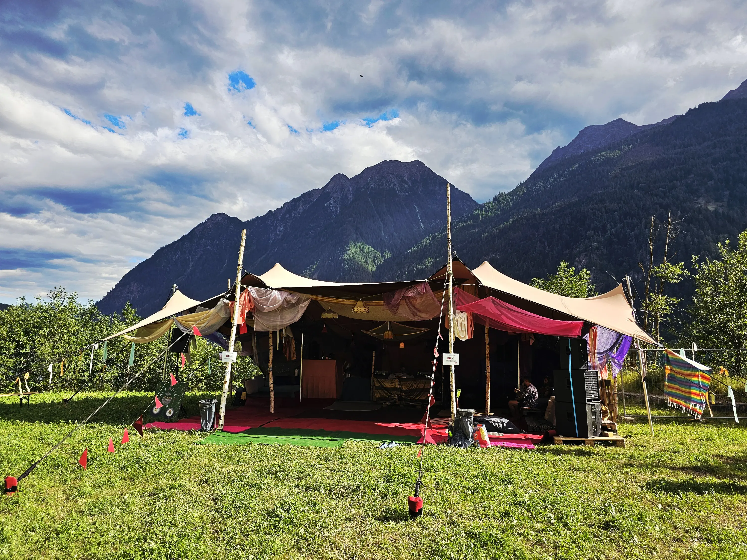Tenda colorata su un prato verde, circondata da montagne, con decorazioni appese e un'atmosfera accogliente. Cielo parzialmente nuvoloso.