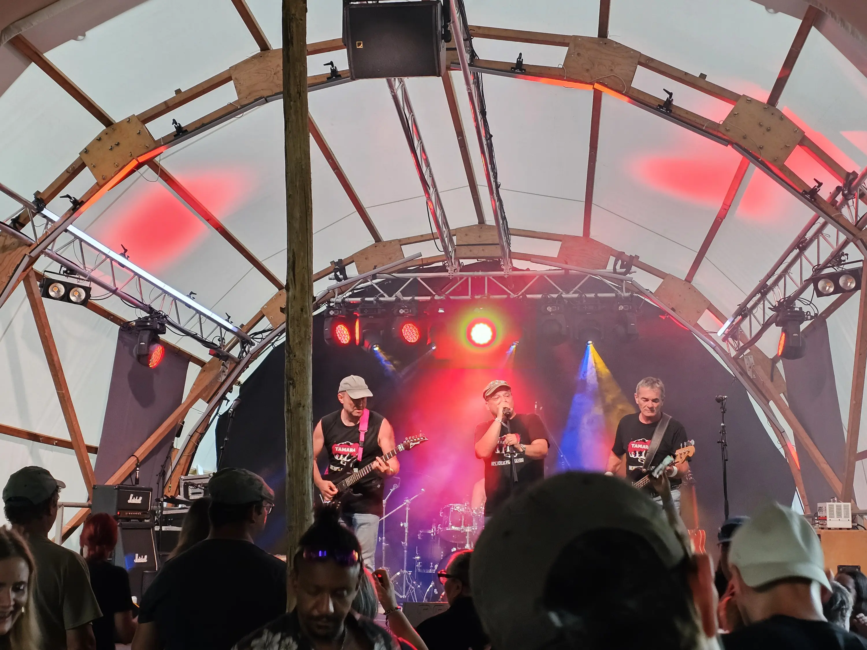 Tre musicisti su un palco all'aperto, con strumenti elettrici, mentre si esibiscono. Illuminaizoni rosse e gialle creano un'atmosfera vivace. Il pubblico è visibile in primo piano, alcuni indossano cappellini.