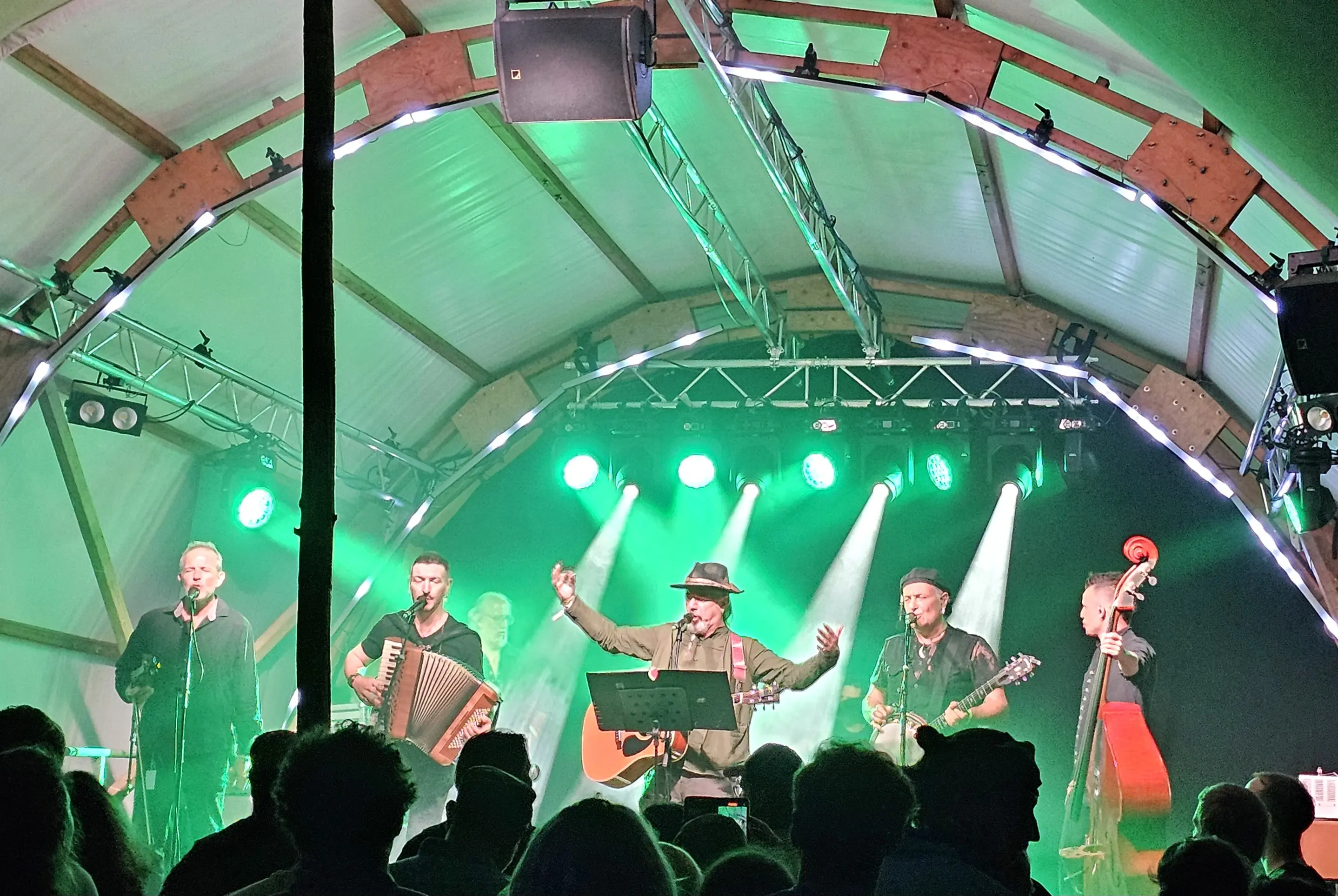Gruppo musicale sul palco in un ambiente illuminato da luci verdi. Cinque musicisti suonano strumenti come chitarre e fisarmoniche, mentre il cantante principale si esibisce al centro. Il pubblico è visibile in primo piano, immerso nella performance.