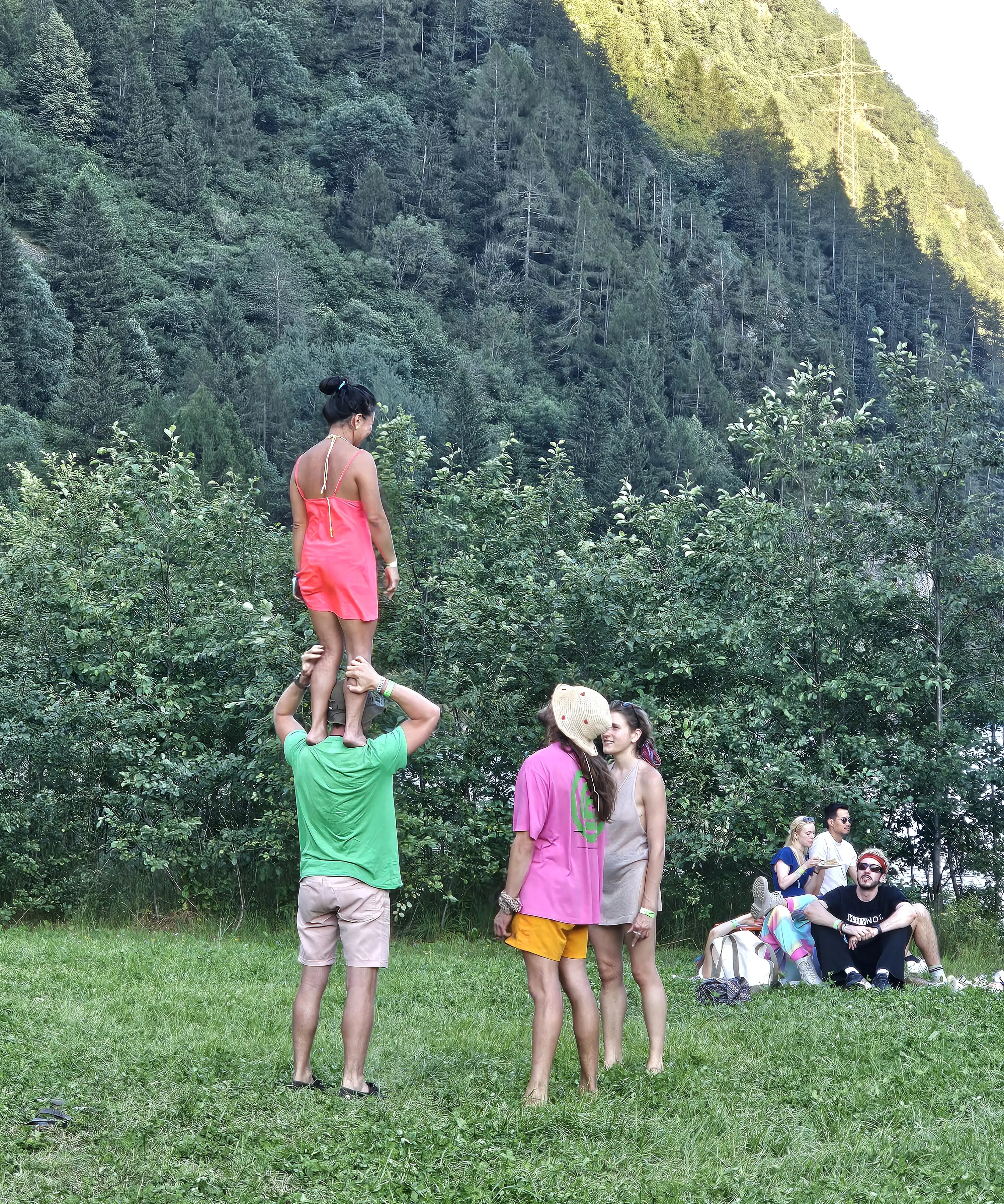 Un gruppo di persone in un campo verde, con una persona che sostiene un'altra in cima alle spalle. Sullo sfondo ci sono alberi e montagne. Altri partecipanti stanno seduti a terra, sorridendo e guardando.