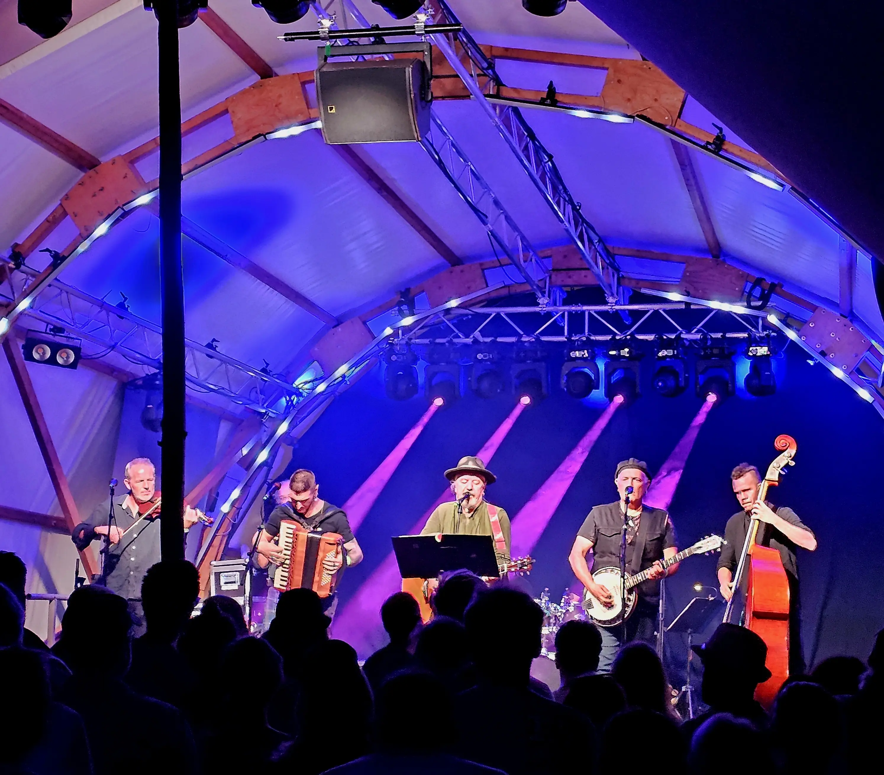 Una band che suona sul palco, con diversi musicisti accompagnati dai loro strumenti. La scena è illuminata da luci blu e viola, creando un'atmosfera vivace mentre il pubblico ascolta.