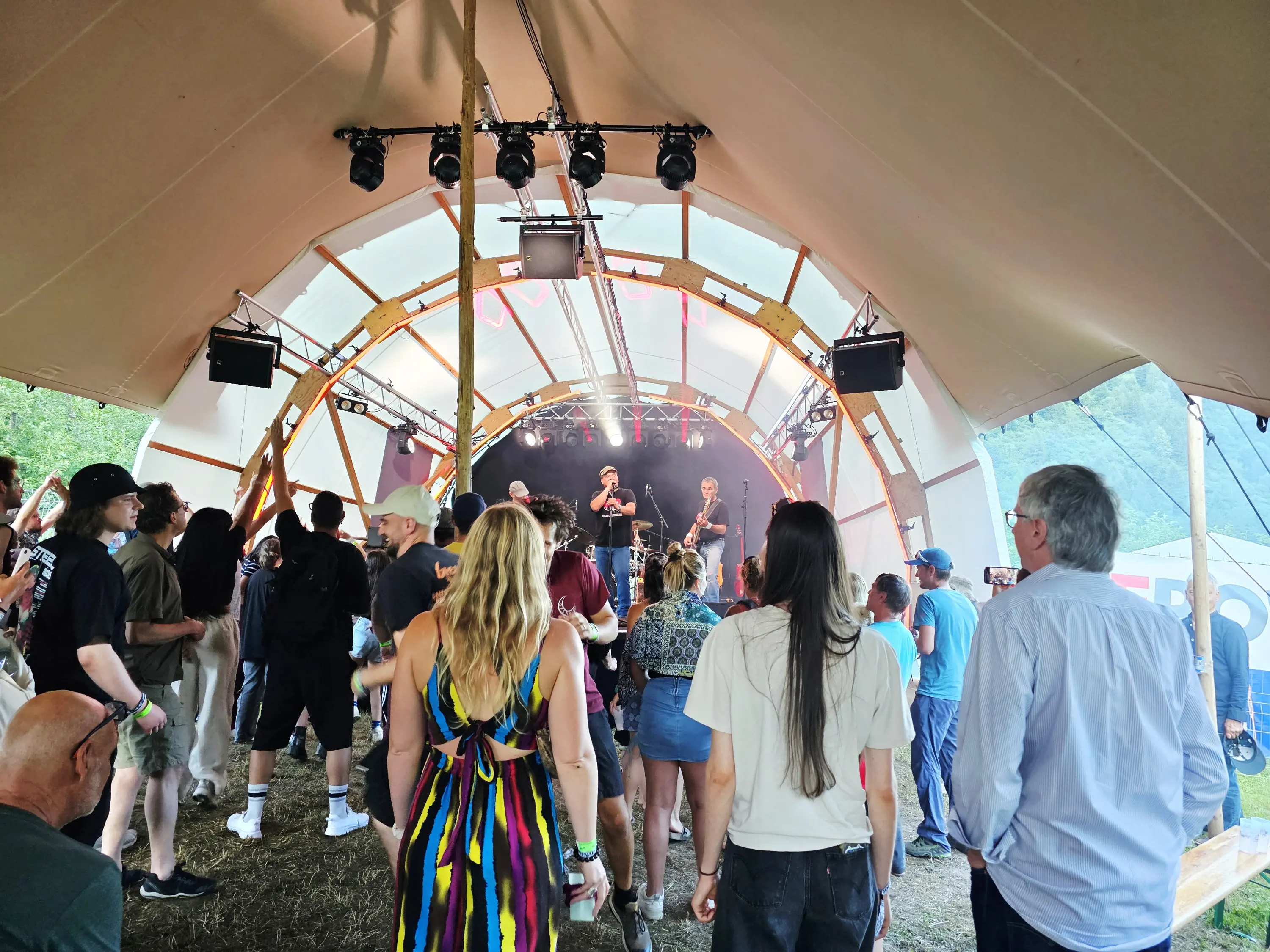 Una folla di persone balla e si diverte sotto una tenda, mentre una band suona sul palco. Le luci si accendono e si spengono, creando un'atmosfera vivace e festosa.