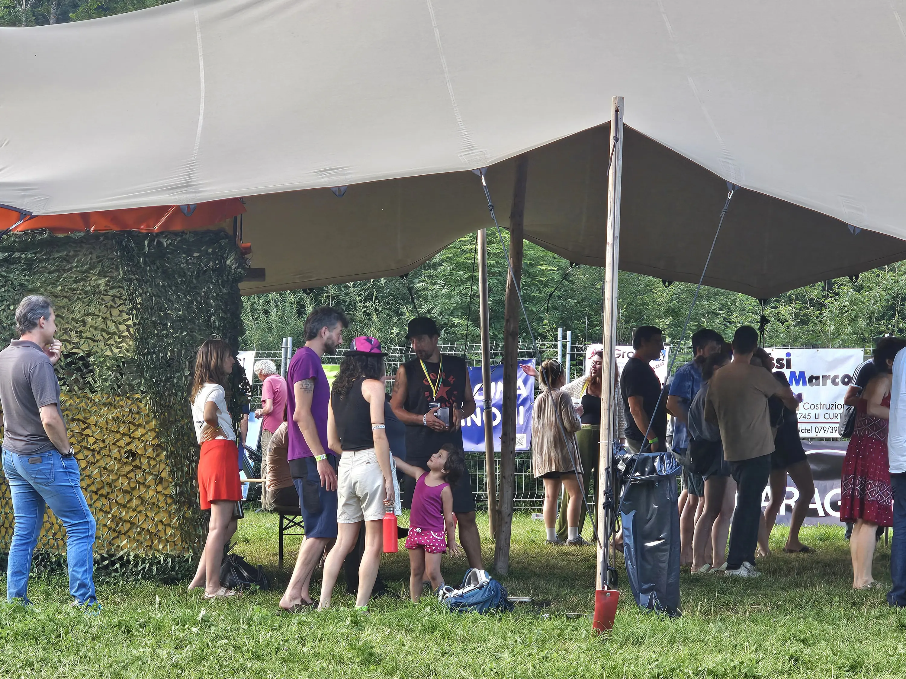 Gruppo di persone sotto una tenda, alcune in fila mentre altre chiacchierano. C'è una bambina con un vestito rosa che gioca vicino. Sullo sfondo si vedono poster e banners.